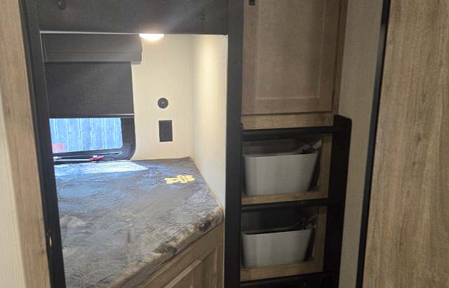 Inside an RV rental, a dark-blanketed bunk bed with a window is on the left, opposite shelves holding white storage bins.. Palomino Puma 2025