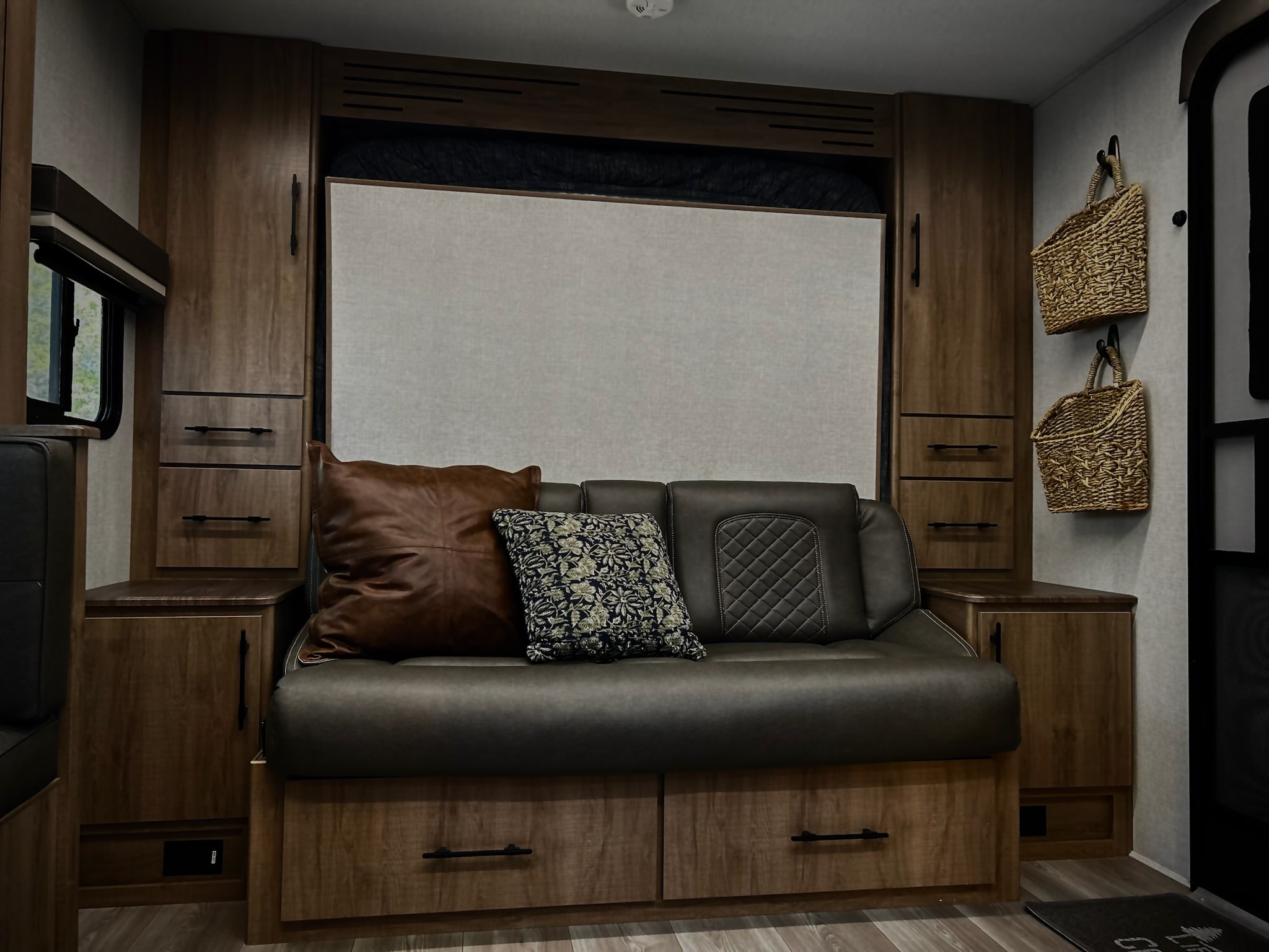 Inside an RV rental: a plush dark sofa with pillows, surrounded by warm wood cabinets and wall-mounted storage baskets.. Grand Design Imagine XLS 2024