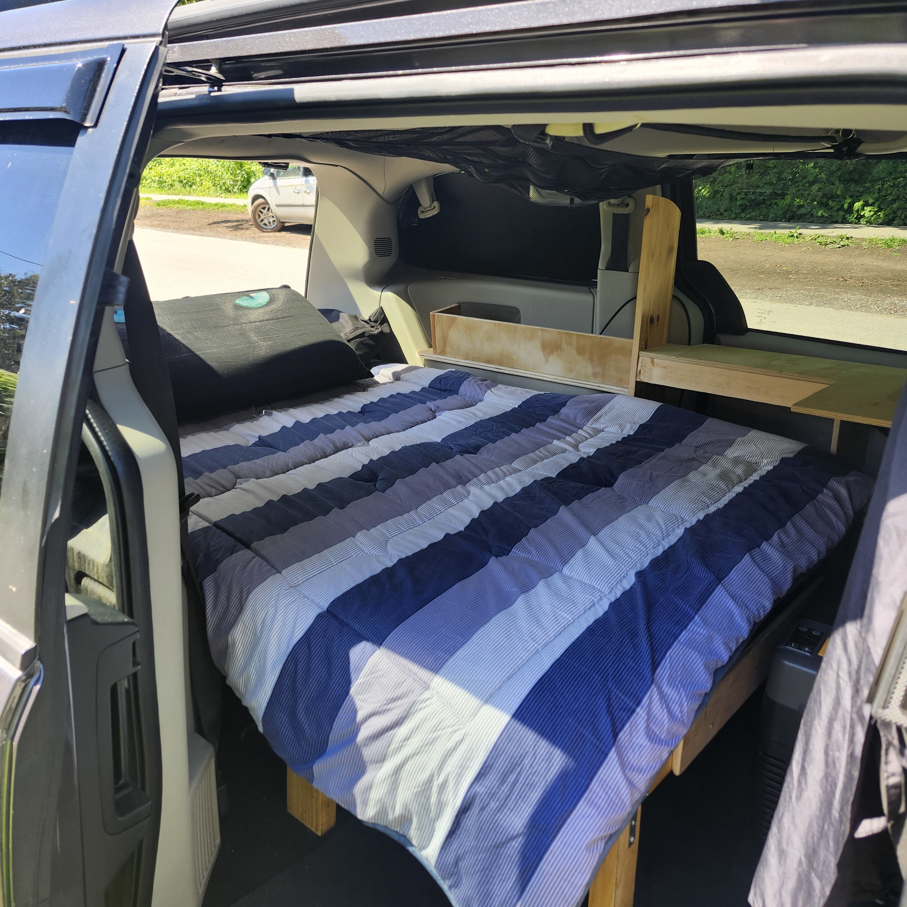 Inside a custom camper van, ideal for RV rental. Features a comfortable bed, pillow, and built-in wooden storage shelves.. Chrysler Dodge Grand Caravan 2015