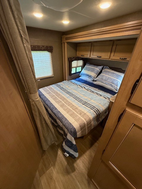 Cozy RV rental bedroom featuring a comfortable bed, striped bedding, overhead cabinets, and a window for natural light.. Thor Freedom Elite 2017