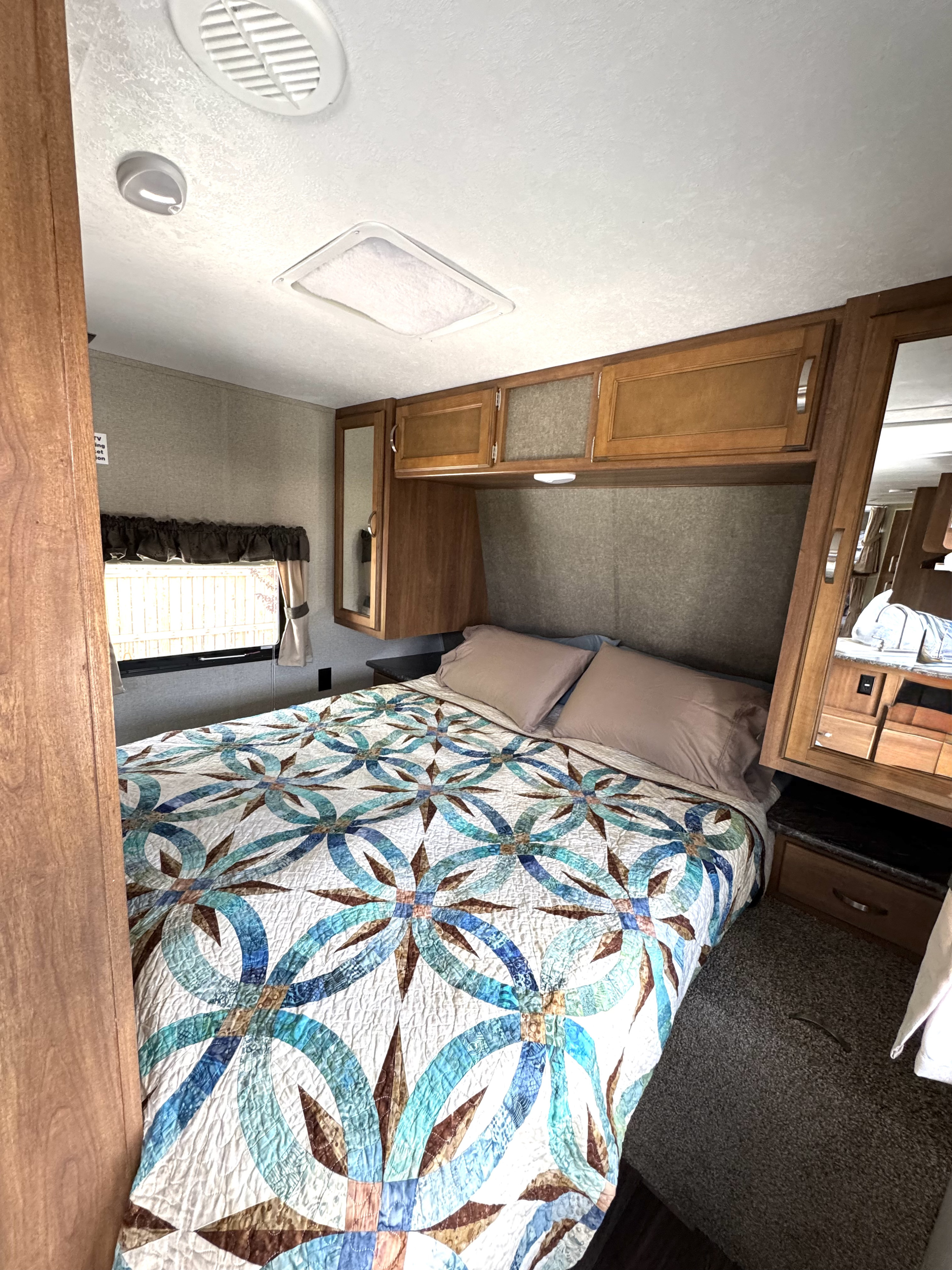 An RV rental bedroom with a blue, teal, and brown patterned quilt on the bed, wooden cabinets, and window.. Keystone RV Springdale SSR 2018