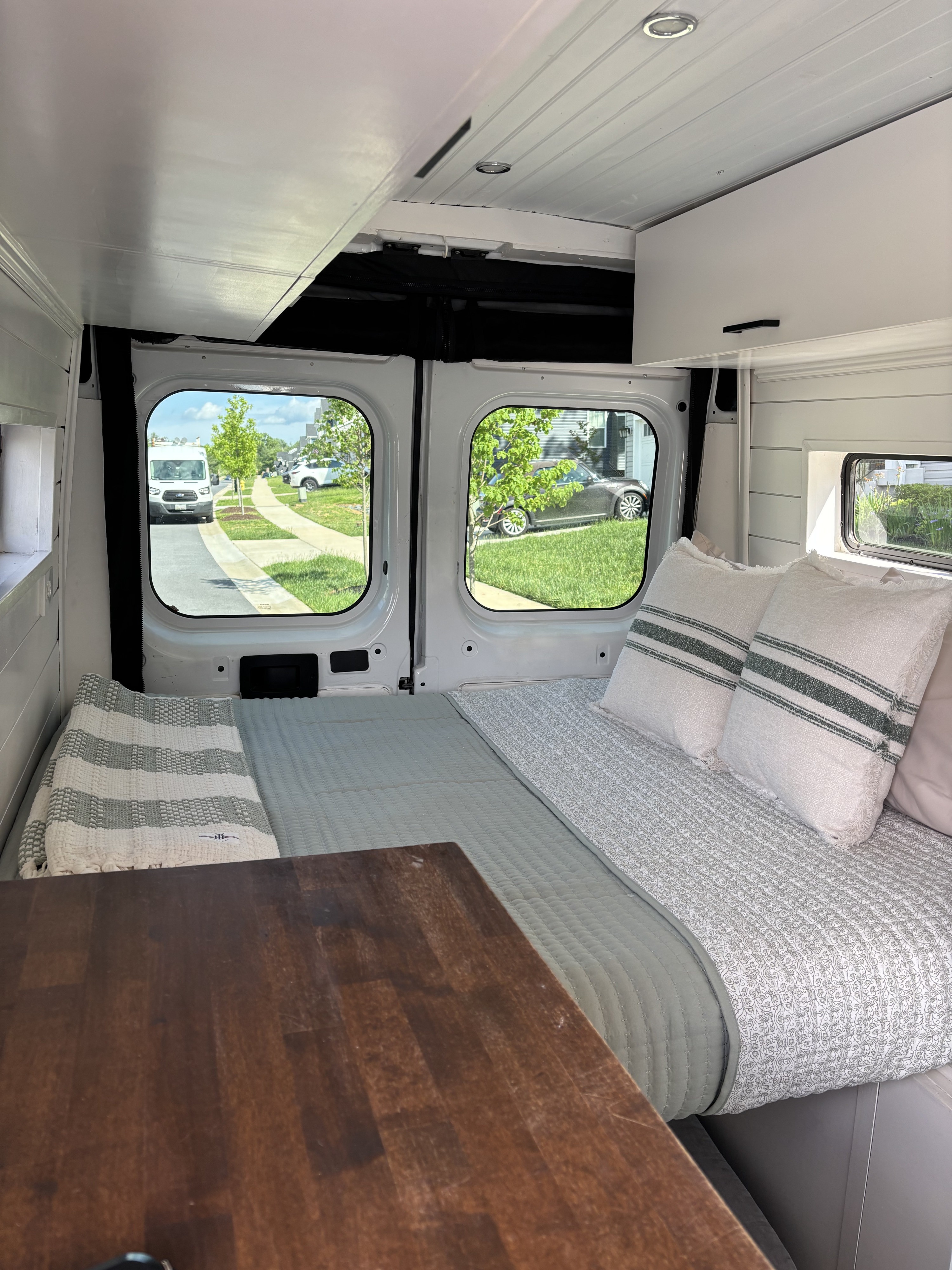 Cozy RV rental interior: bed with pillows, wooden table, and street views through large windows, perfect for travel.. Ram Ram ProMaster 2500 2019