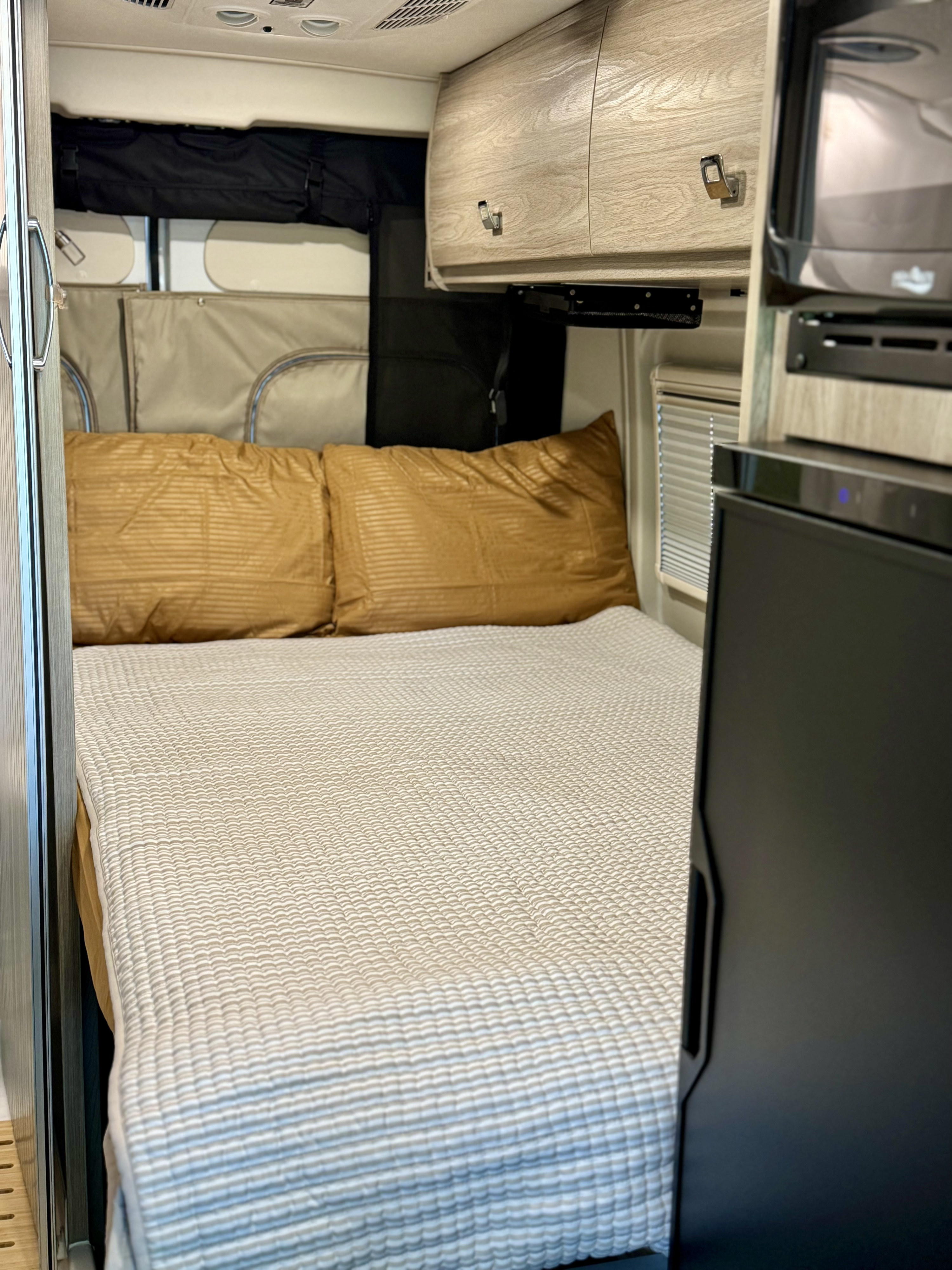 Inside a comfy RV rental, featuring a bed with gold pillows, a quilted comforter, and a built-in fridge.. Winnebago Travato 2023