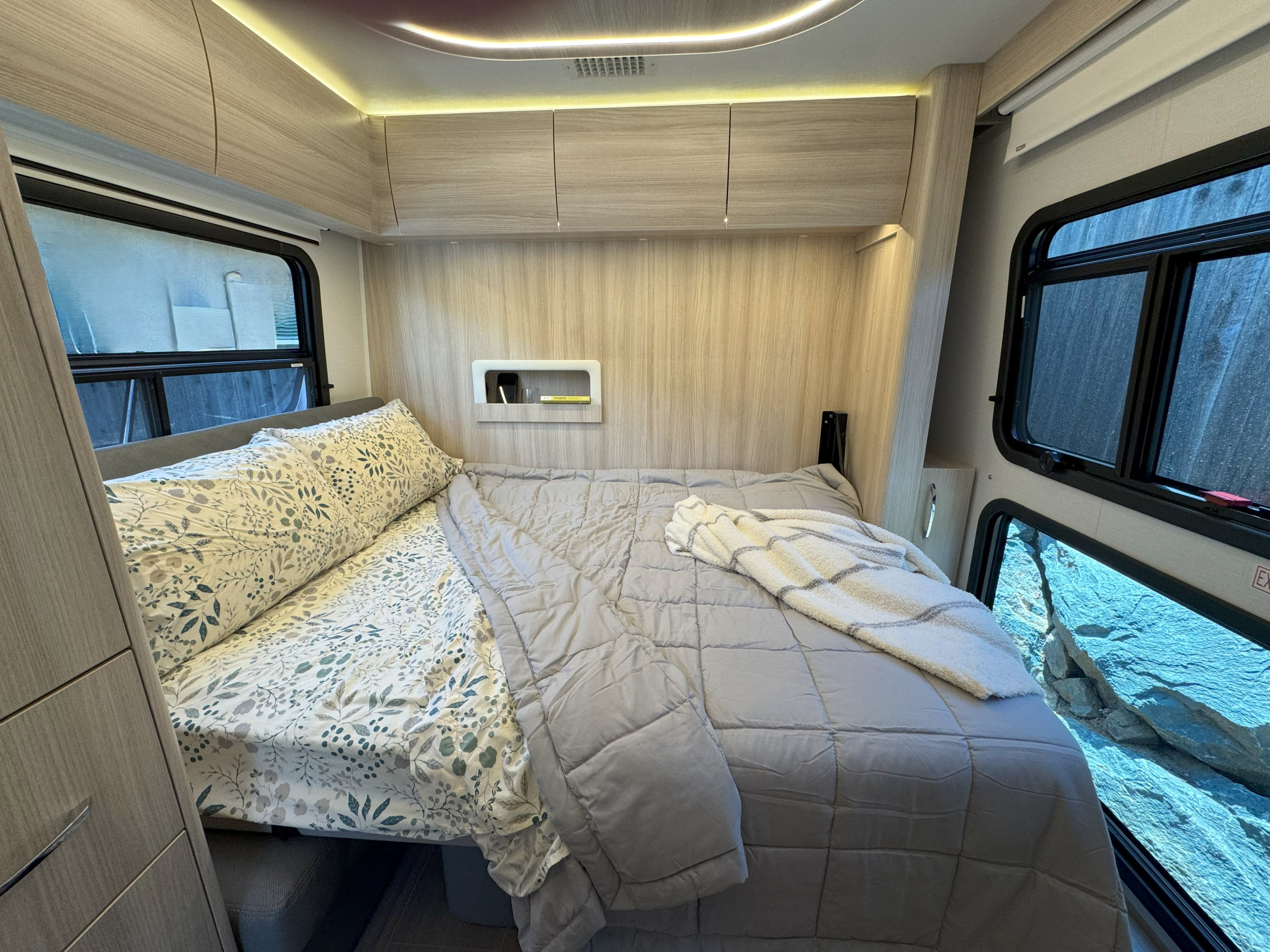 Cozy RV rental bedroom featuring a bed with patterned sheets, light wood decor, and windows offering scenic views.. Leisure Travel Vans Unity 2022
