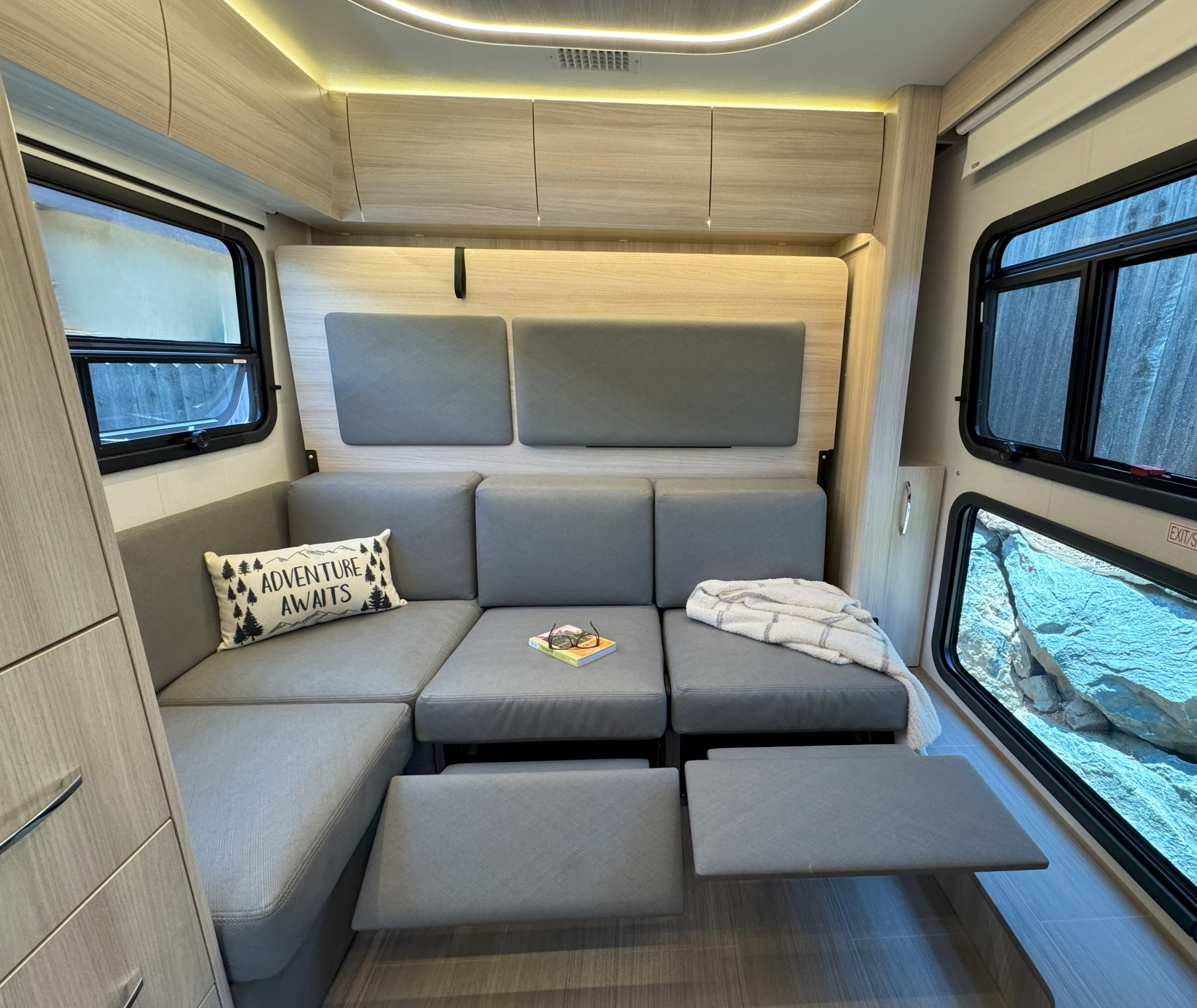 RV rental interior: A modern and inviting lounge with versatile grey sofa, pull-out footrests, overhead storage, and scenic windows.. Leisure Travel Vans Unity 2022