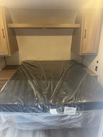 New, plastic-wrapped bed in RV rental bedroom, with overhead storage cabinets and a shelf.. Keystone RV CM25B25 2025