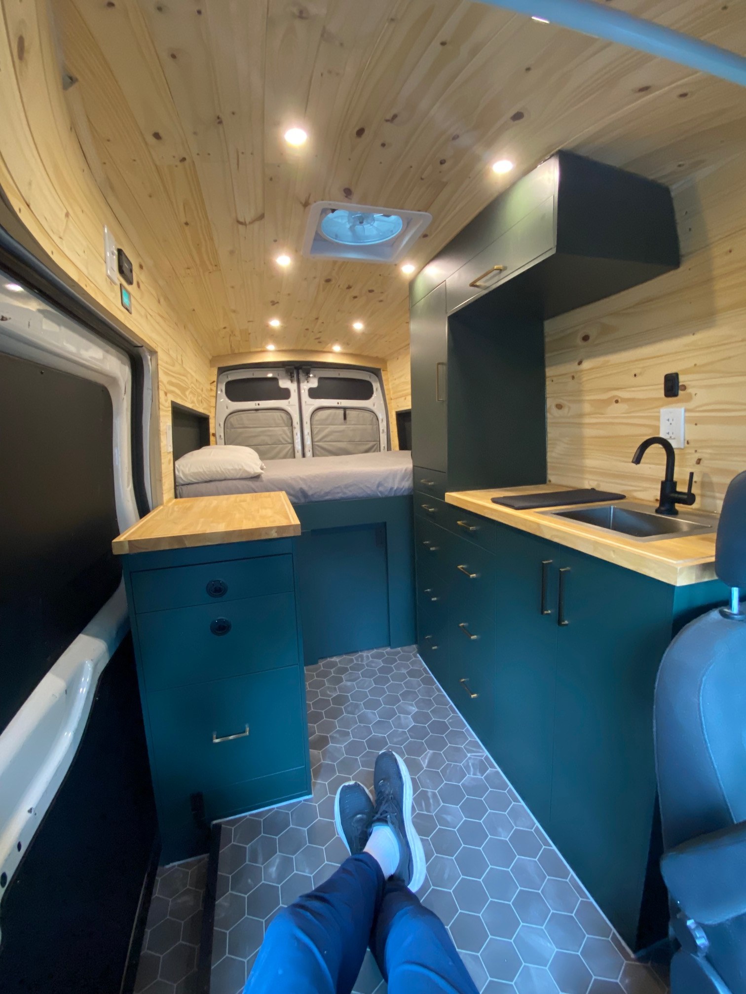 Modern RV rental interior with wood-paneled ceiling, teal cabinets, kitchen, and cozy bed. Perfect for van life!. Ford Van 2016