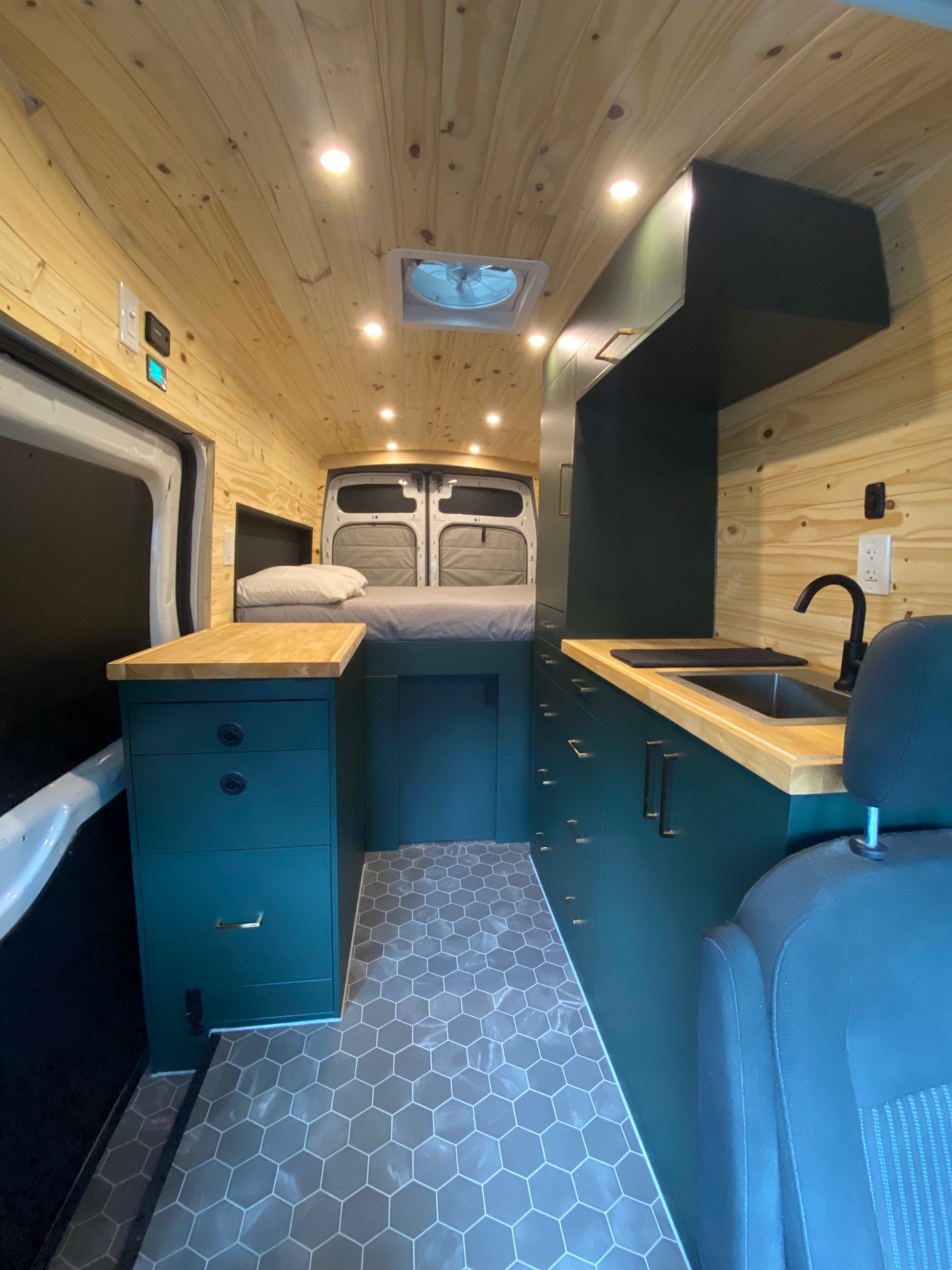 Modern RV rental interior with wood paneling, dark green cabinets, compact kitchen, and cozy bed area.. Ford Van 2016