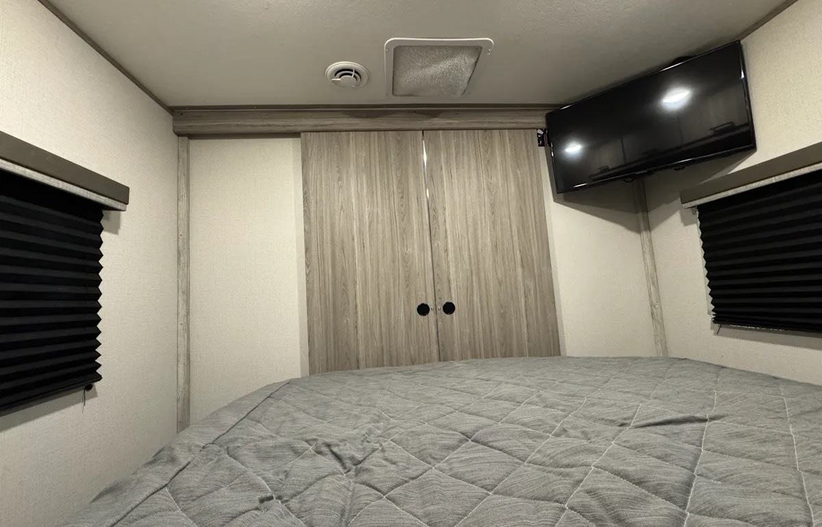 RV rental bedroom interior featuring a comfortable bed, mounted TV, closet doors, and window blinds. Cozy and functional space.. Keystone RV Passport SL 2021