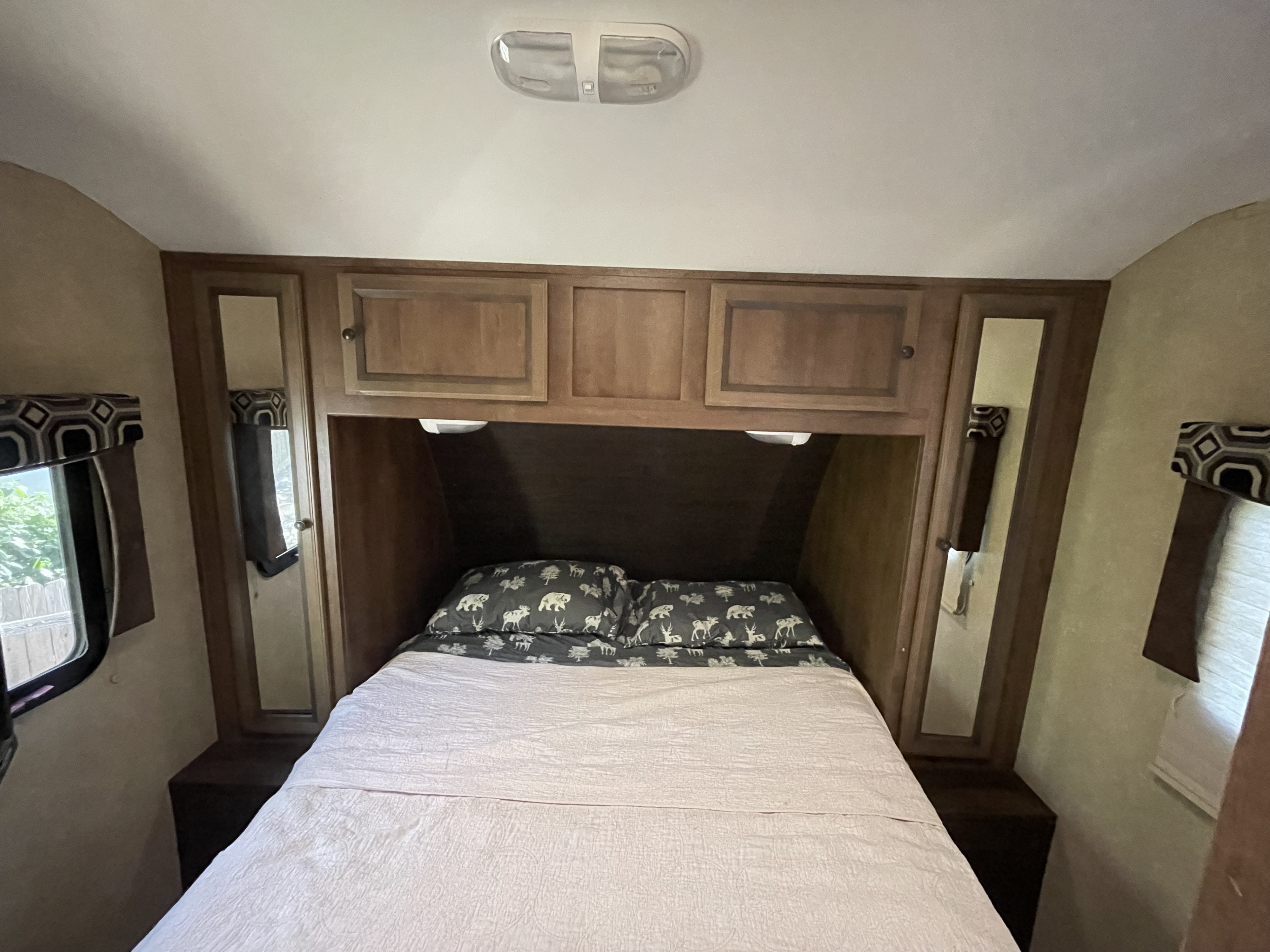 Inside an RV rental: a compact bedroom with a bed, animal-themed pillows, overhead cabinets, and side windows.. Cruiser RV Radiance 2015