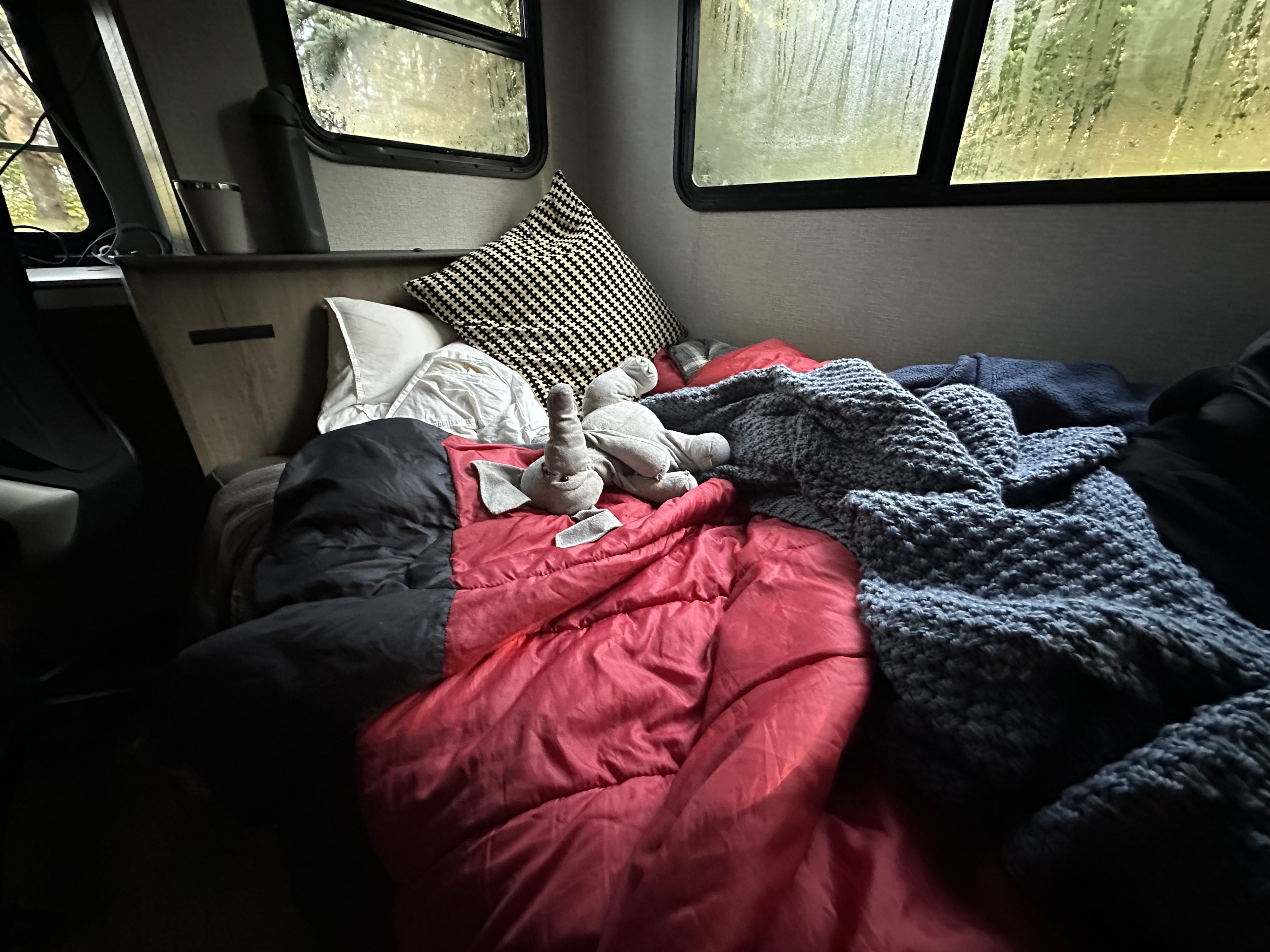 Inside a comfortable RV rental: a bed with red sleeping bag, blue blanket, and elephant toy, looking out at rainy trees.. Grand Design Imagine 2023