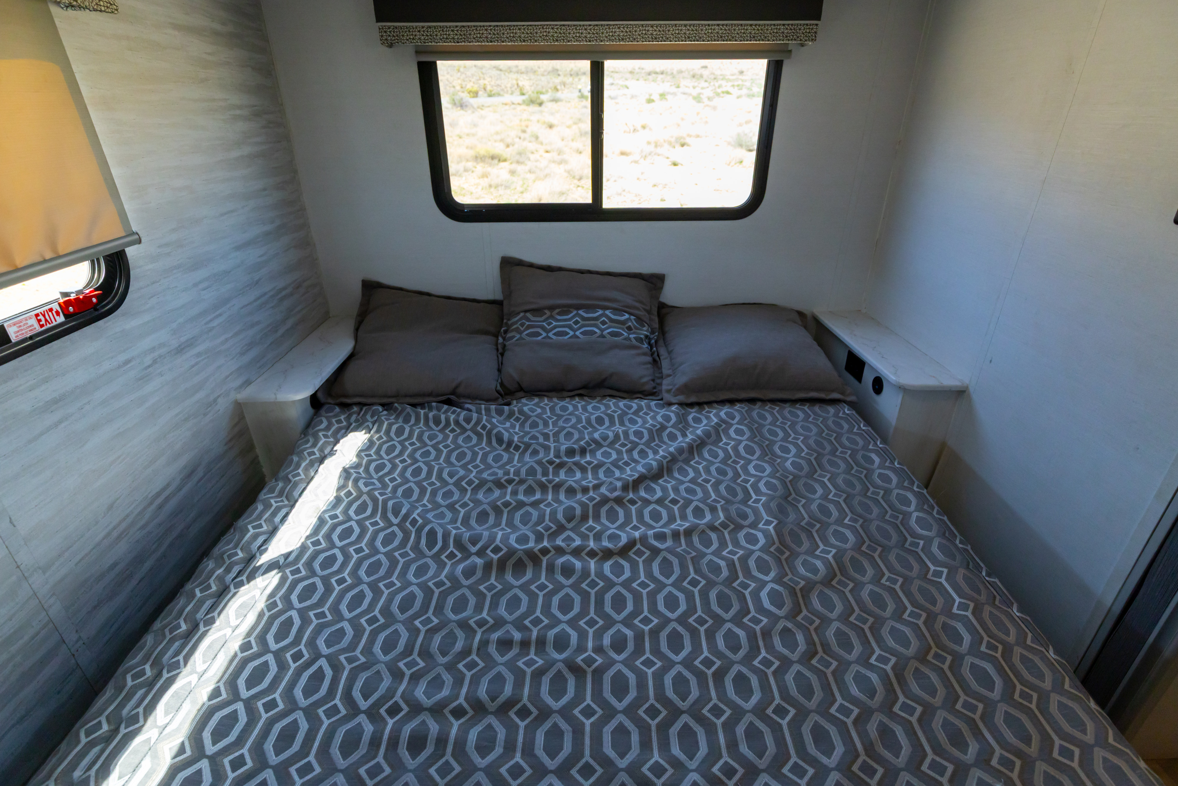 Inside an RV rental: a neatly made bed with grey geometric bedding, pillows, and a window overlooking an arid landscape.. Gulf Stream Conquest 2024