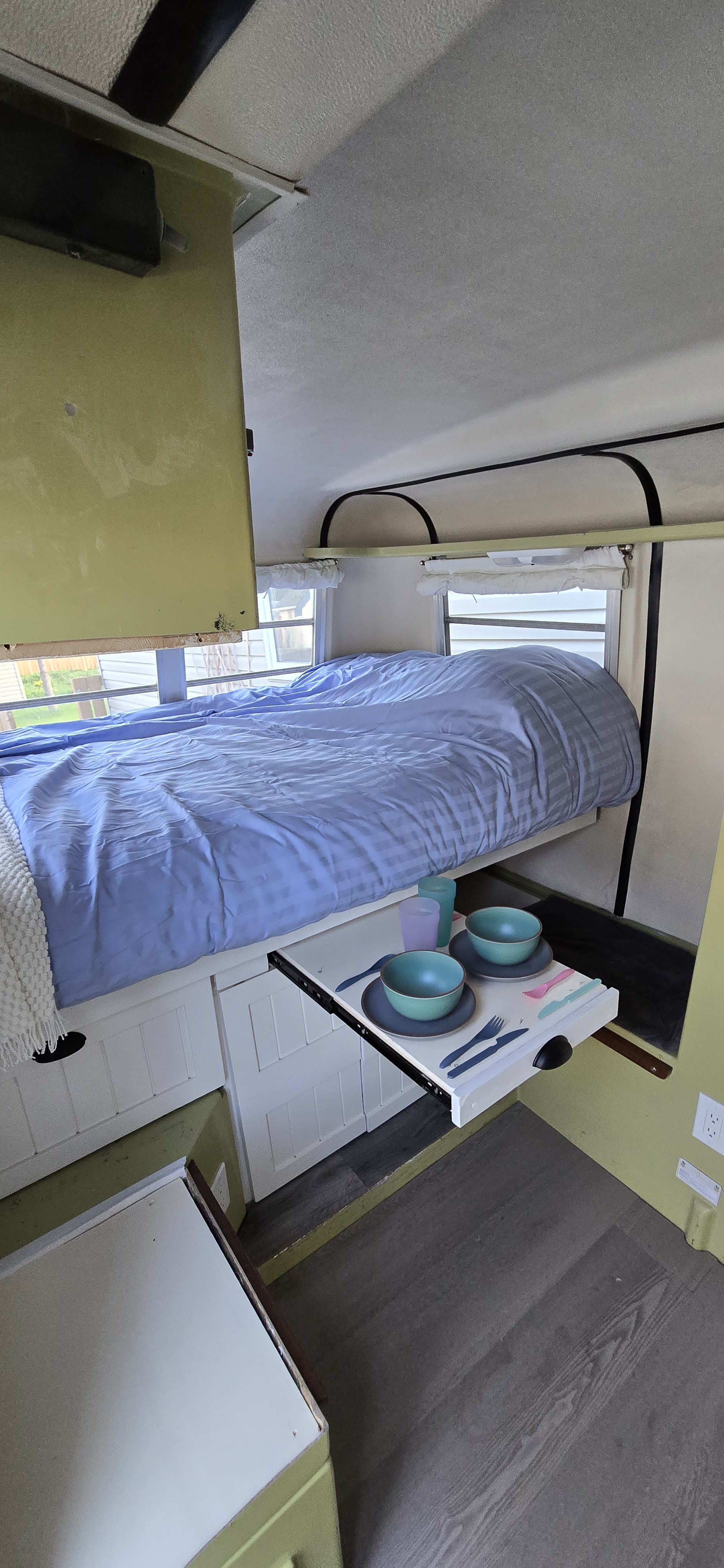 Cozy RV rental interior with a blue-sheeted bed & innovative pull-out dining table set with dishes.. Trillium Rv 1300 1973