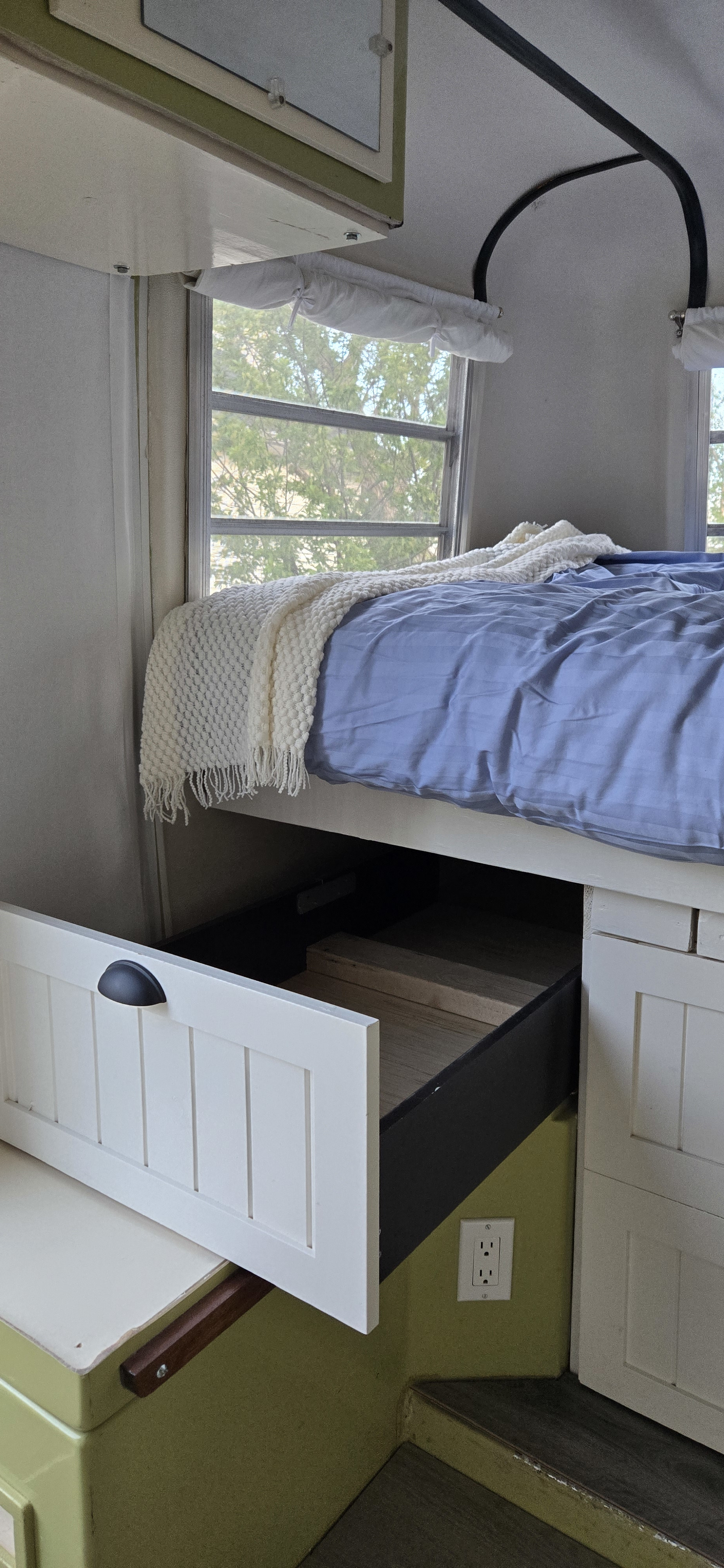 Inside a cozy RV rental: a bed with blue sheets, a white blanket, and an open under-bed storage drawer revealing wooden blocks.. Trillium Rv 1300 1973