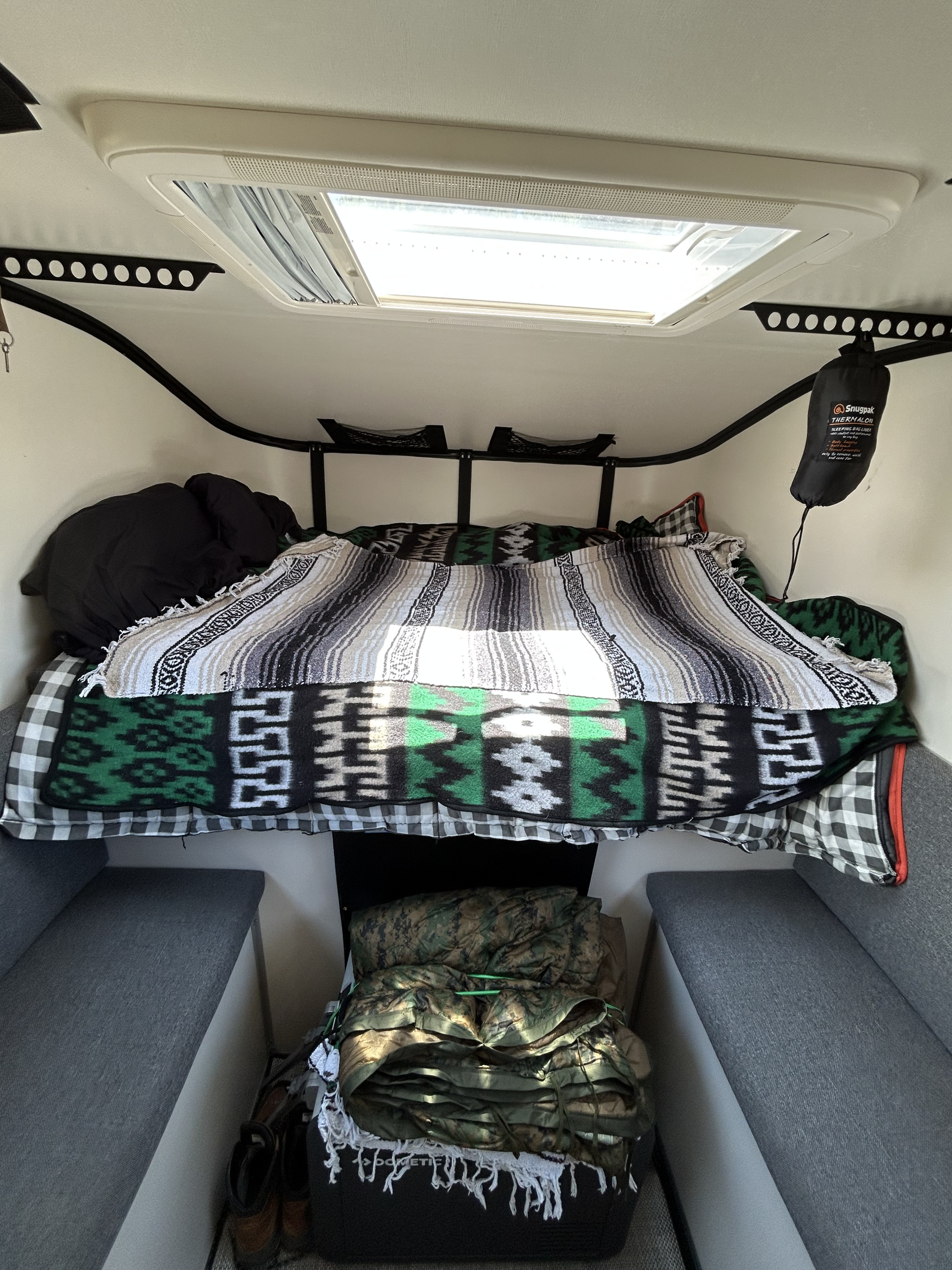 Cozy RV rental interior featuring a bed with patterned blankets, skylight, and compact storage. Ideal for adventure travel.. Toyota Tacoma 2018