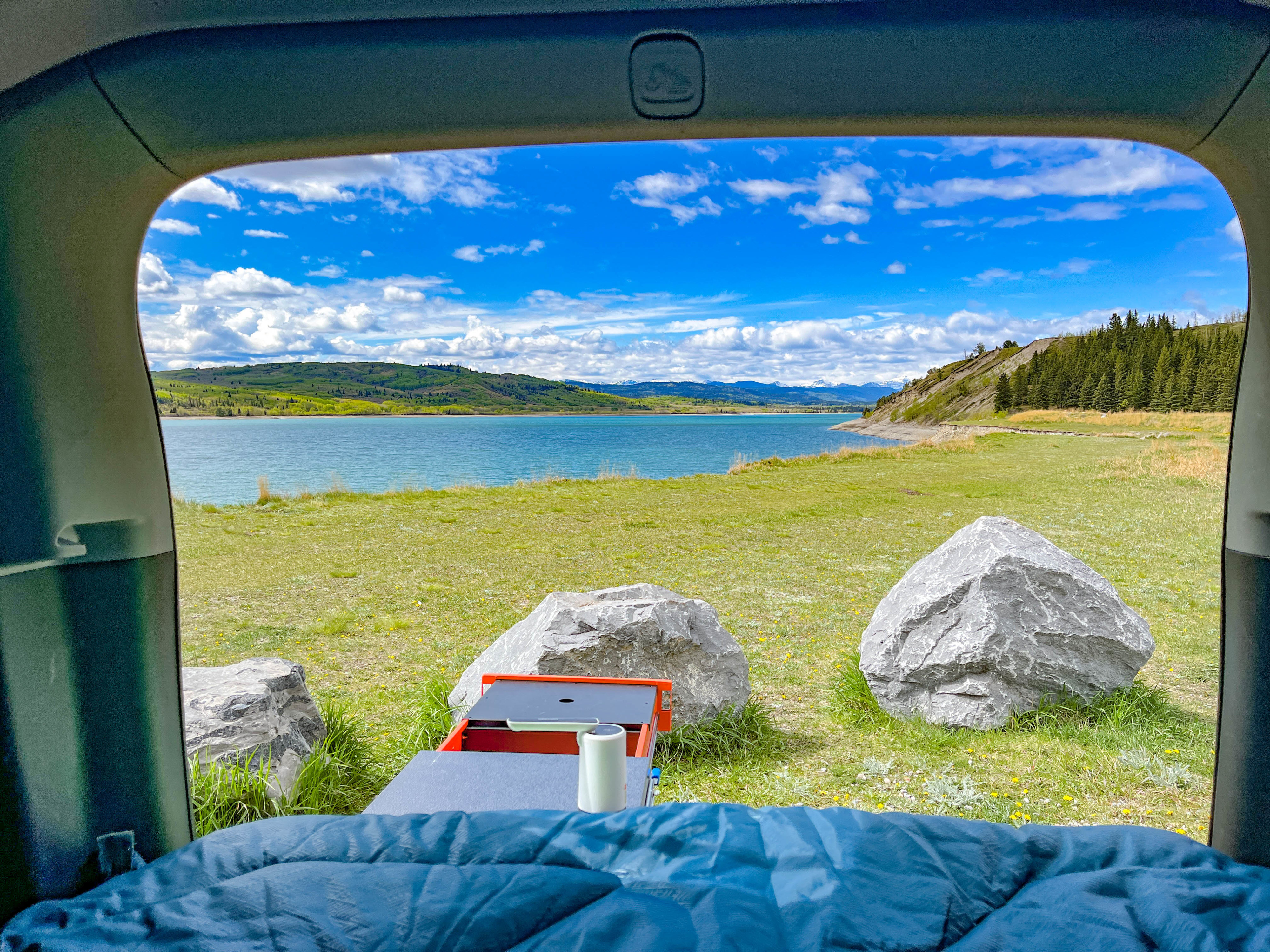 Inside an RV rental, a comfy bed faces a stunning lake and mountain landscape under a bright sky. Outdoor adventure awaits.. honda 2019 CRV Red 2019