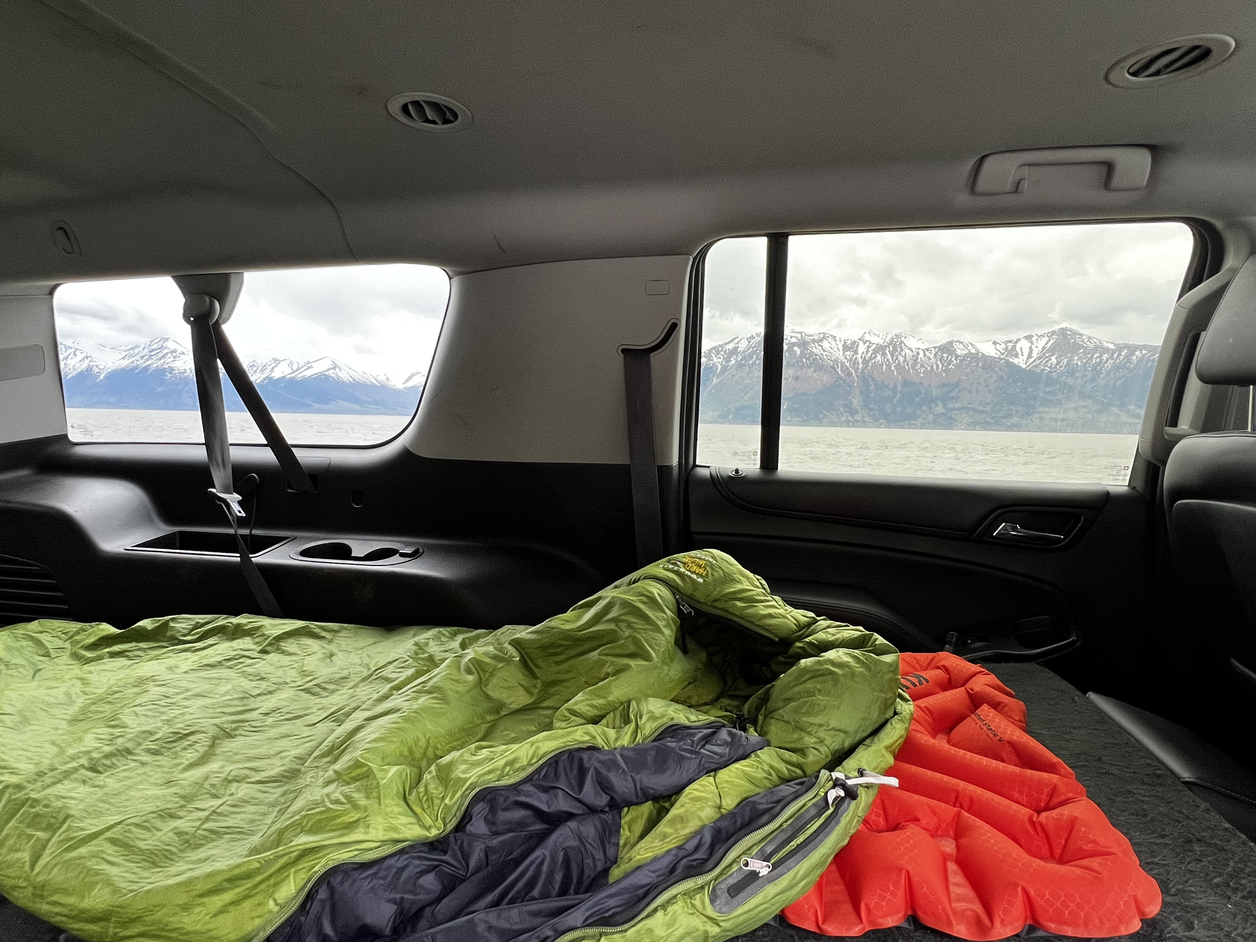 Car camping setup inside a vehicle with sleeping bag & pad, offering scenic views of snow-capped mountains & water. Ideal for an RV rental adventure.. Chevrolet Suburban 2015