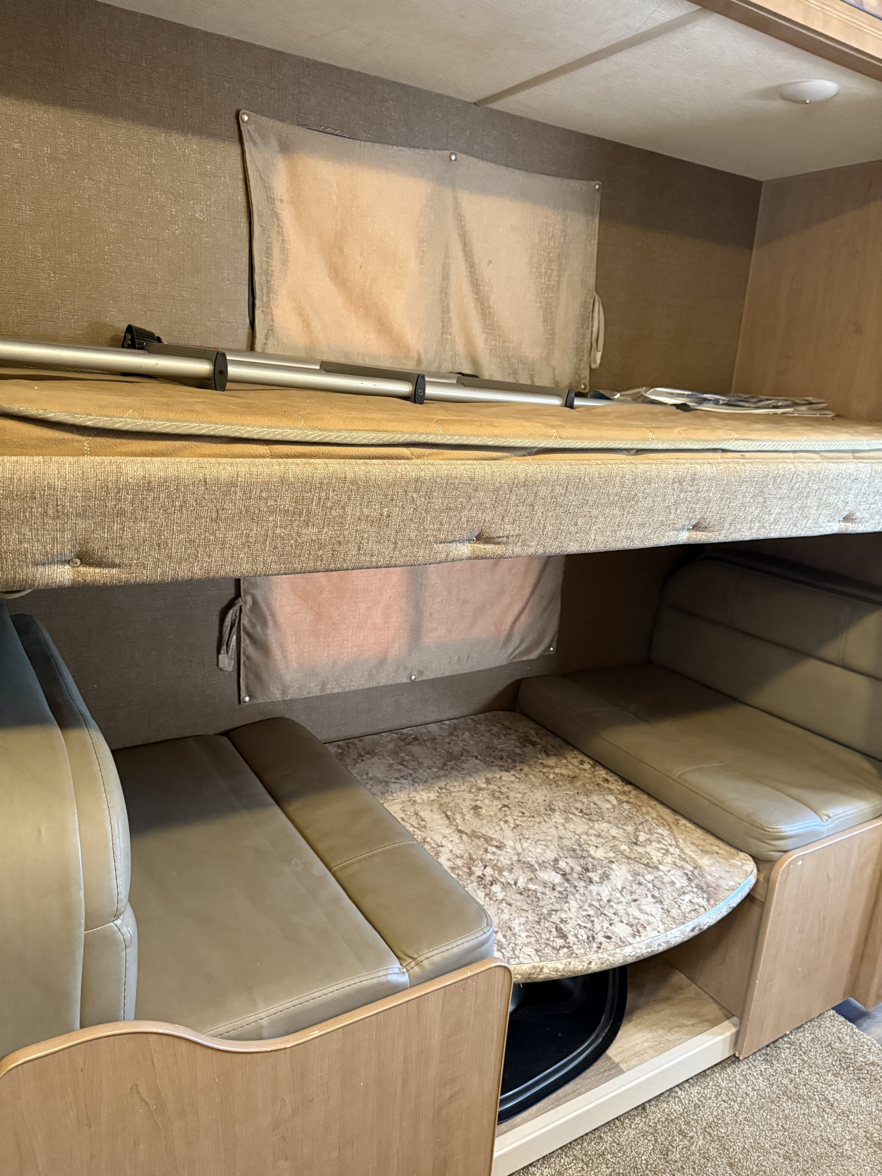 An RV rental interior reveals a top bunk bed & lower dinette area with tan upholstered seating and a patterned table.. Thor Four Winds 2018
