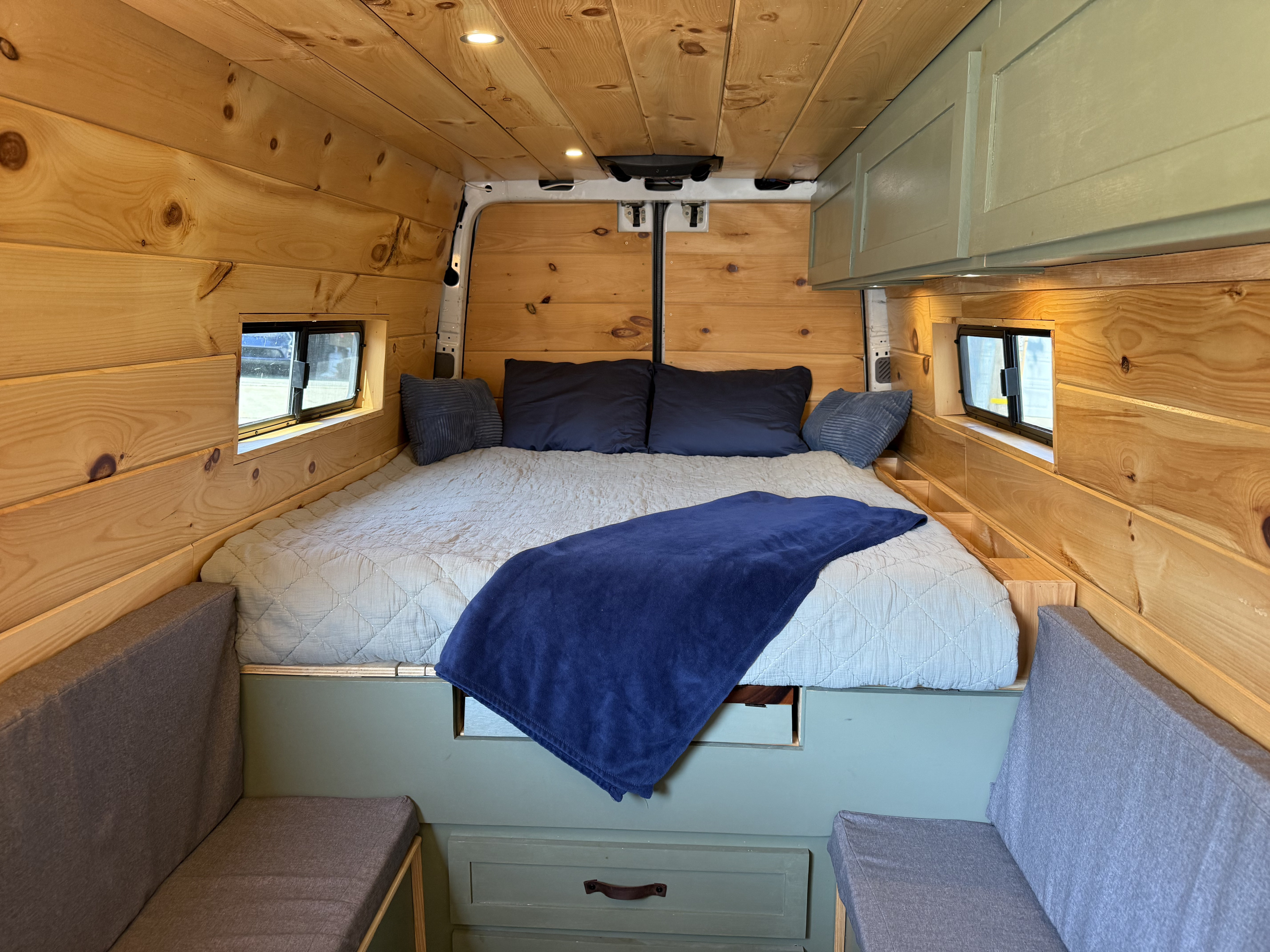 Cozy RV rental interior with a comfortable bed, warm wooden walls, and sage green cabinetry for a perfect camper van experience.. Dodge Sprinter 3500 2007