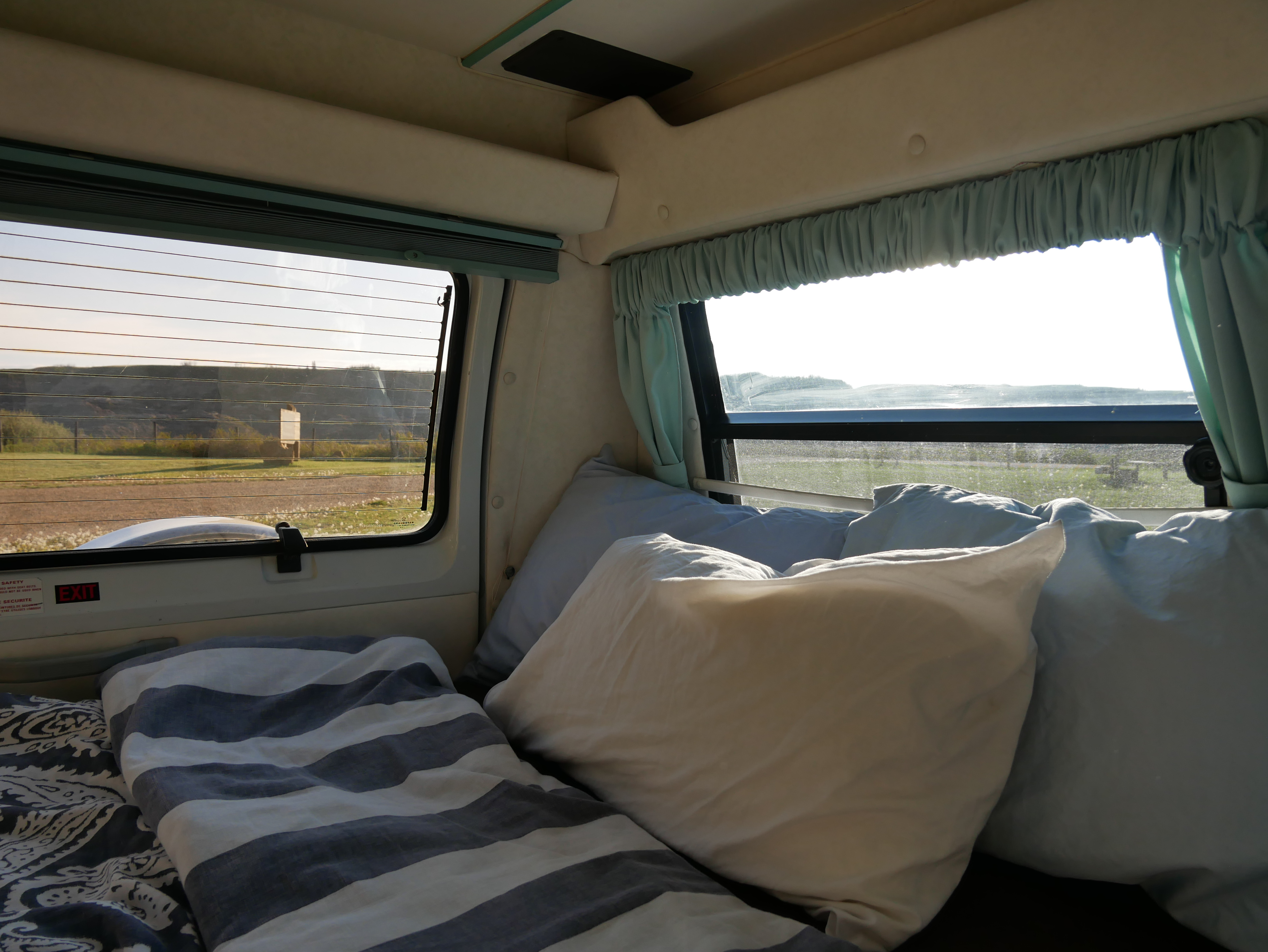 Cozy RV rental interior: bed with striped duvet, pillows, and bright window views of grassy hills.. Leisure Travel Freedom 1994