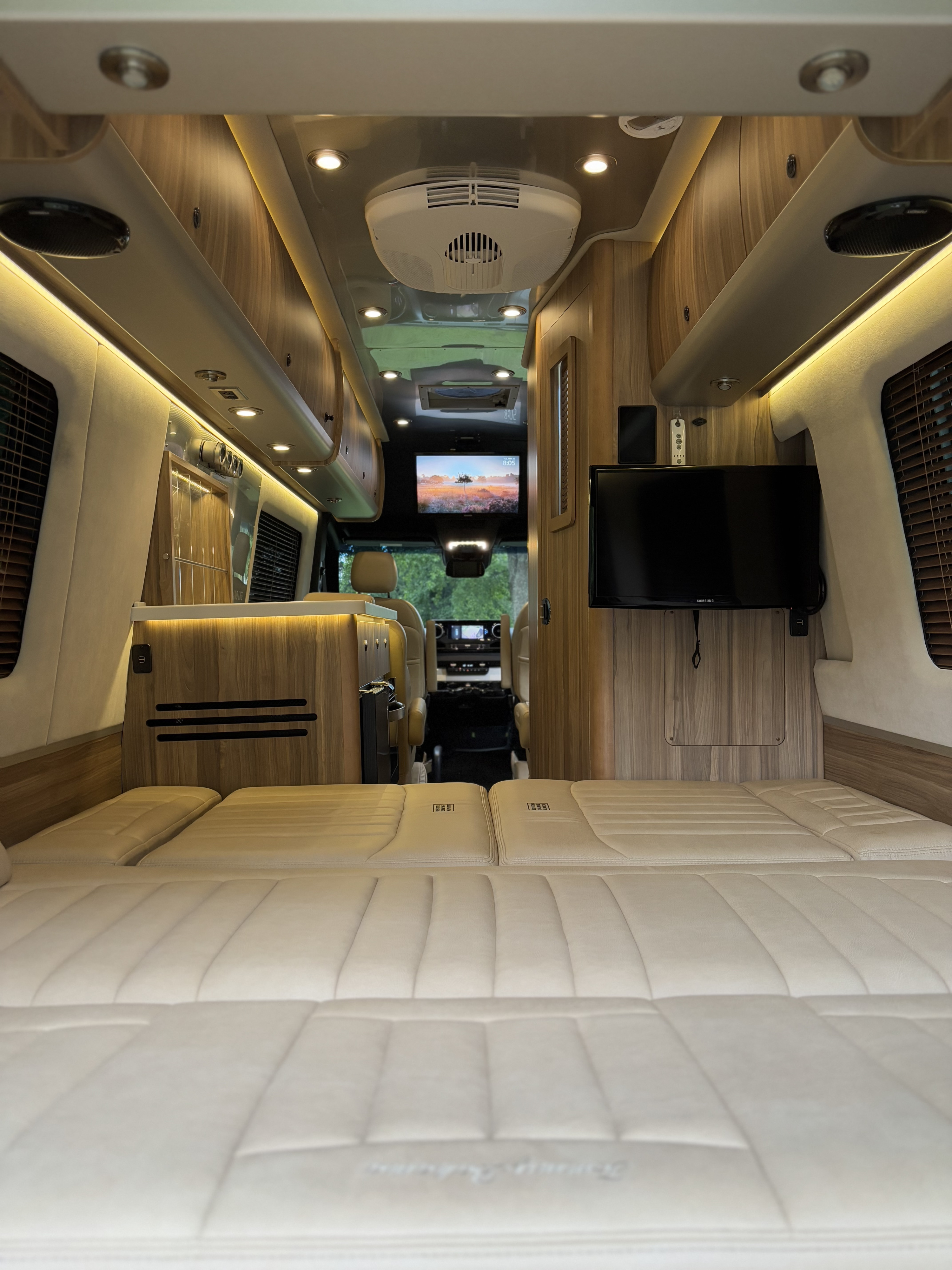 Inside a luxurious RV rental, looking forward. Features a comfortable bed, warm lighting, and wood cabinetry.. Airstream Interstate 2020