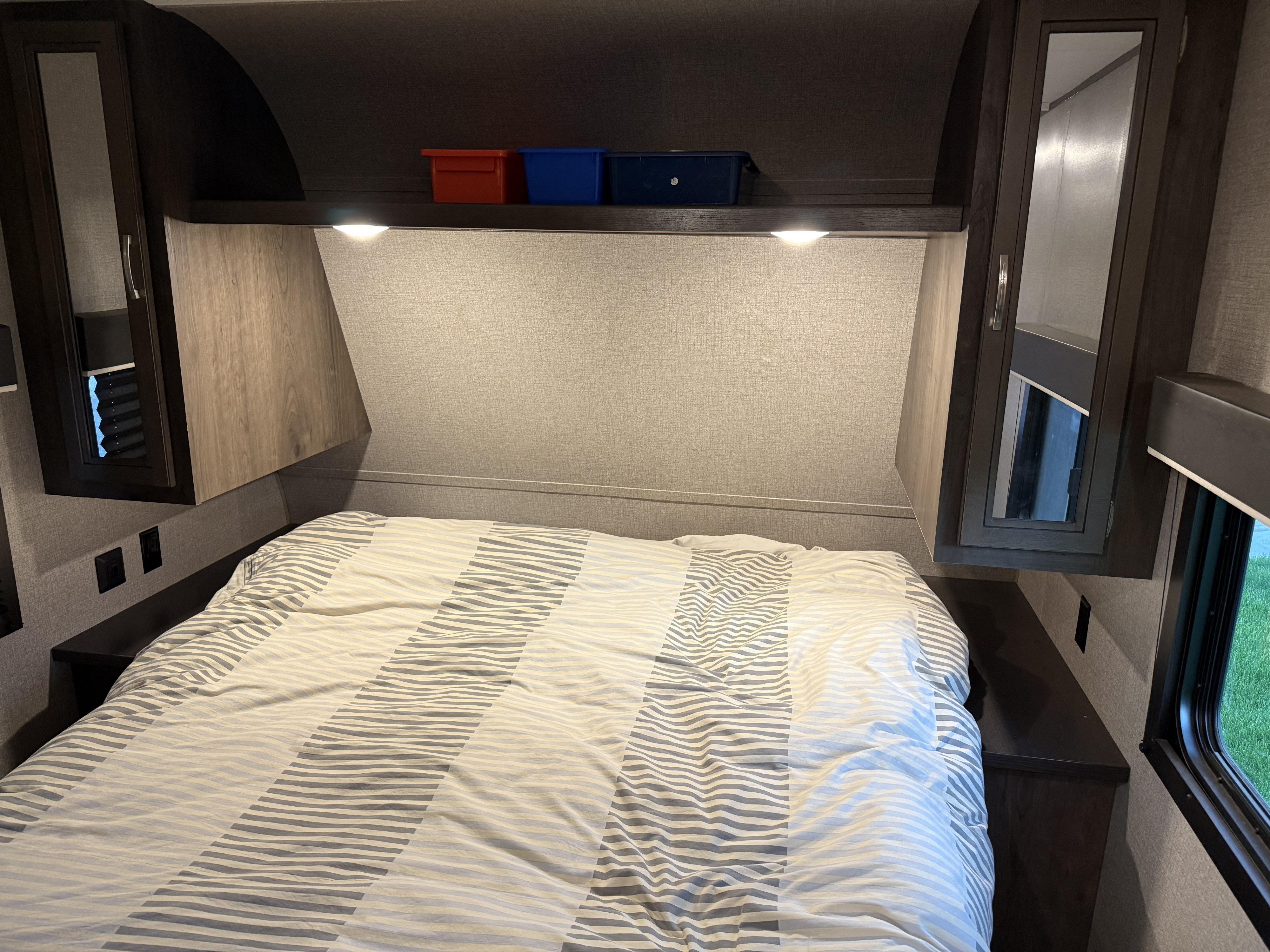 Cozy RV rental bedroom showing striped bedding, overhead storage cabinets, and reading lights above a shelf with bins.. Jayco Jay Flight 2020