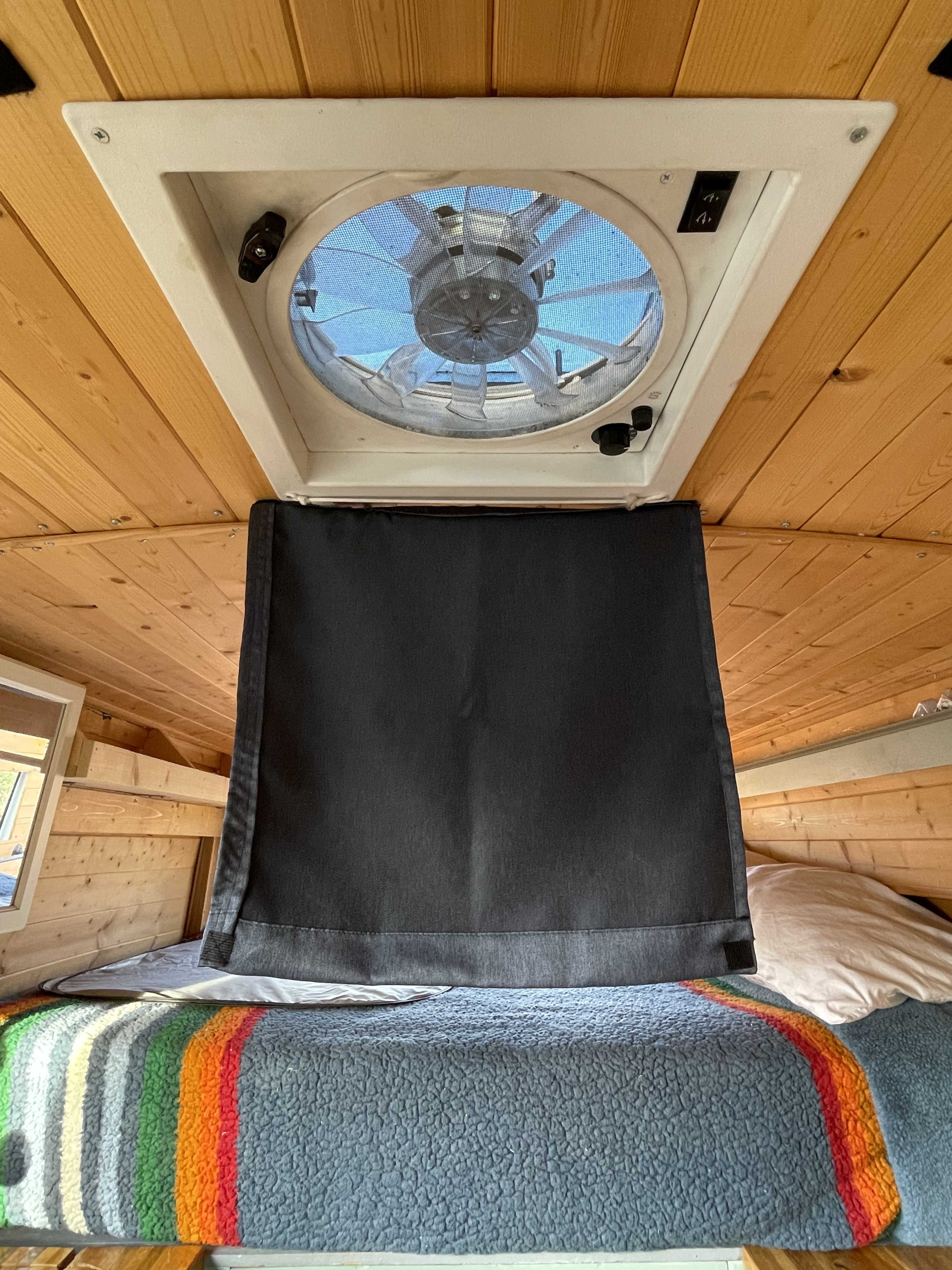 Inside an RV rental: Wooden ceiling, roof vent with fan & screen. Dark curtain covers the bed with a striped blanket.. Ford Transit 2017