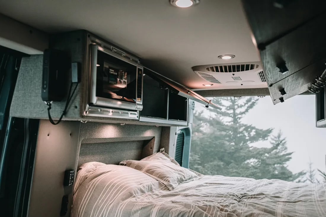 Cozy RV rental interior: comfortable bed, integrated microwave, and overhead cabinets. Misty forest visible outside.. Storyteller Overland Storyteller Overland Class B 2021