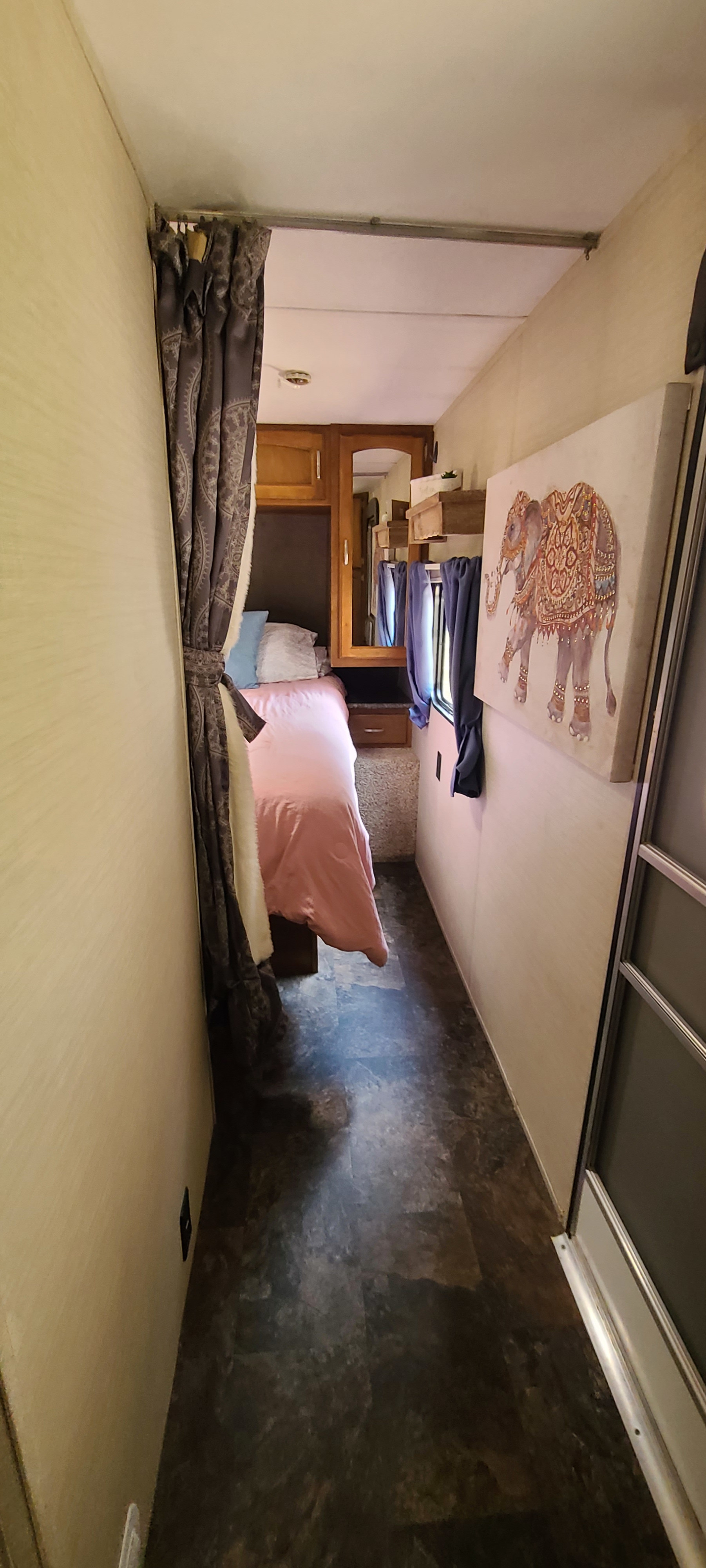 Inside an RV rental: a narrow hallway leads to a compact bedroom with pink bedding, wooden cabinets, and elephant art.. Keystone RV Springdale 2015