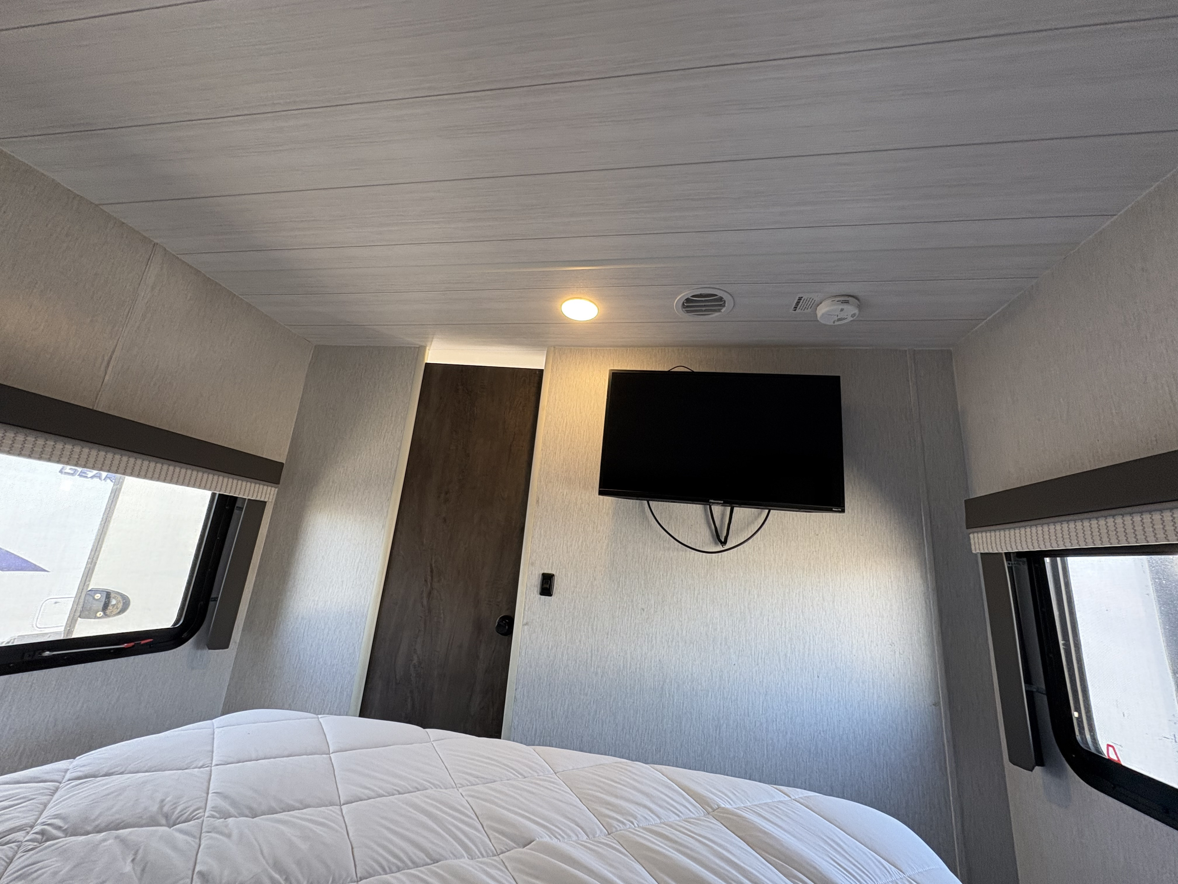 Inside an RV rental: a bright bedroom with a white comforter, wall-mounted TV, side windows, and a dark wood door.. Forest River Wildwood X-Lite 2022