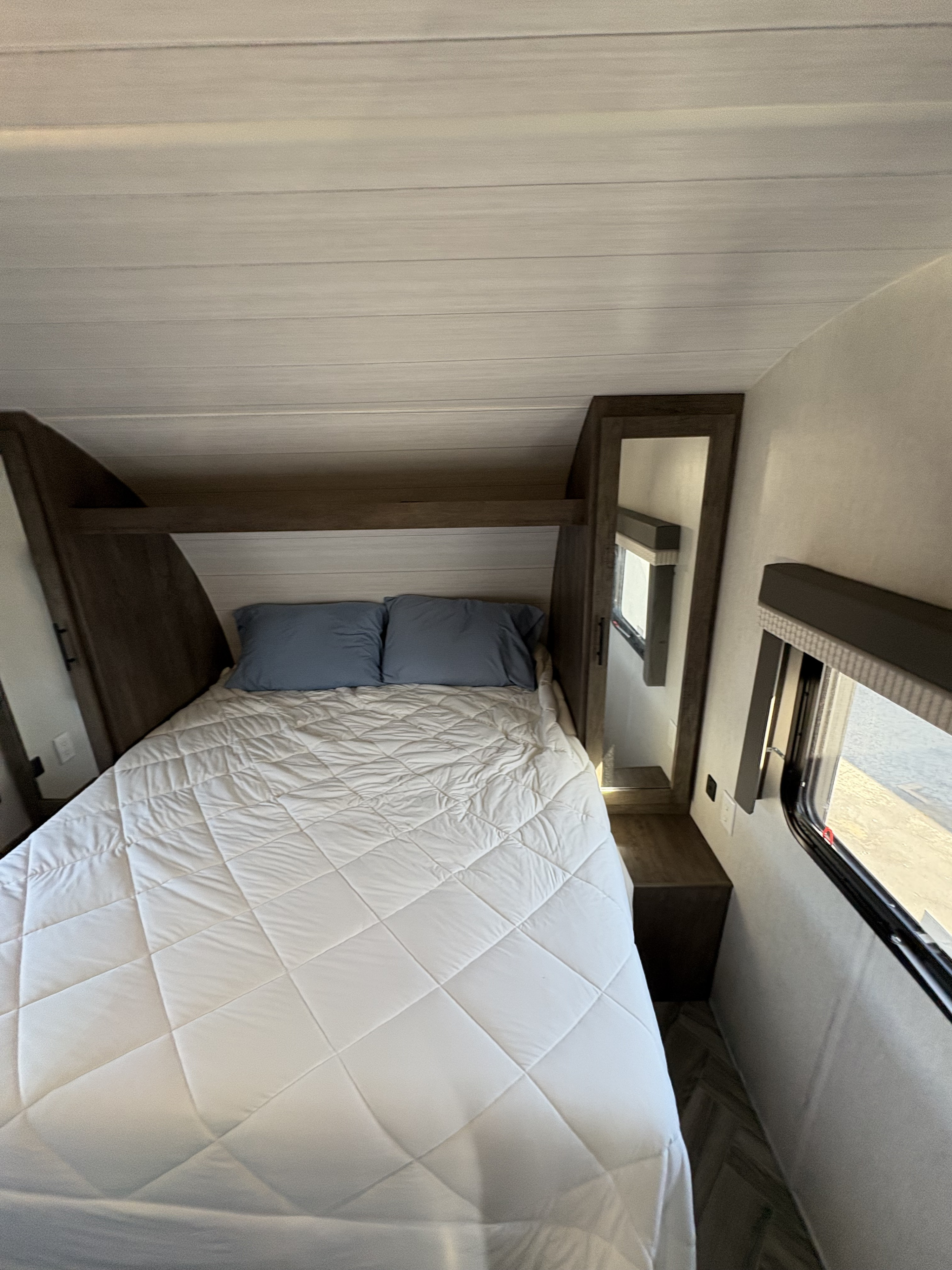 Cozy RV rental bedroom with a white quilted bed, blue pillows, built-in cabinets, and a window for natural light.. Forest River Wildwood X-Lite 2022