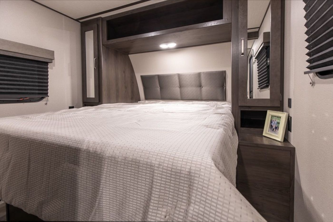 Cozy RV rental bedroom with a large bed, padded headboard, overhead cabinets, and window with blinds.. Grand Design Transcend Xplor 2023