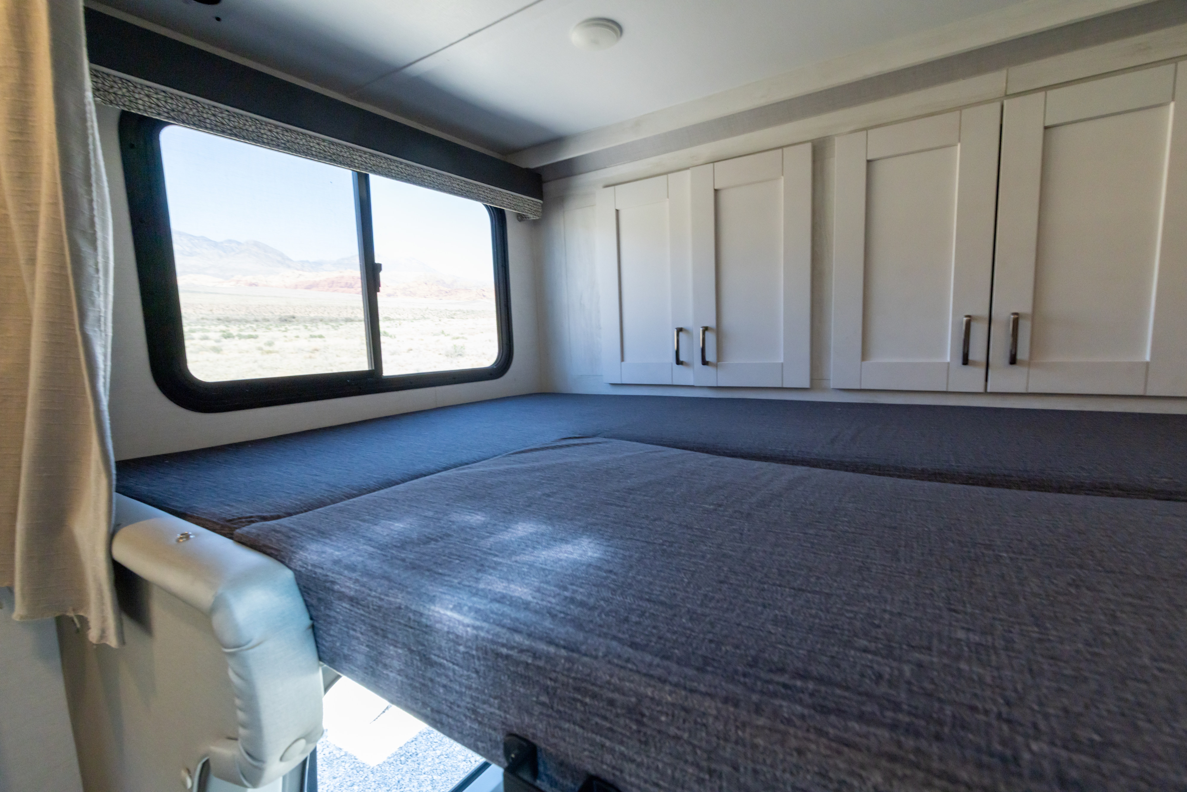Inside an RV rental: A comfortable sleeping area with dark grey cushions, white overhead cabinets, and a wide window showing desert mountains.. Gulf Stream Conquest 2024