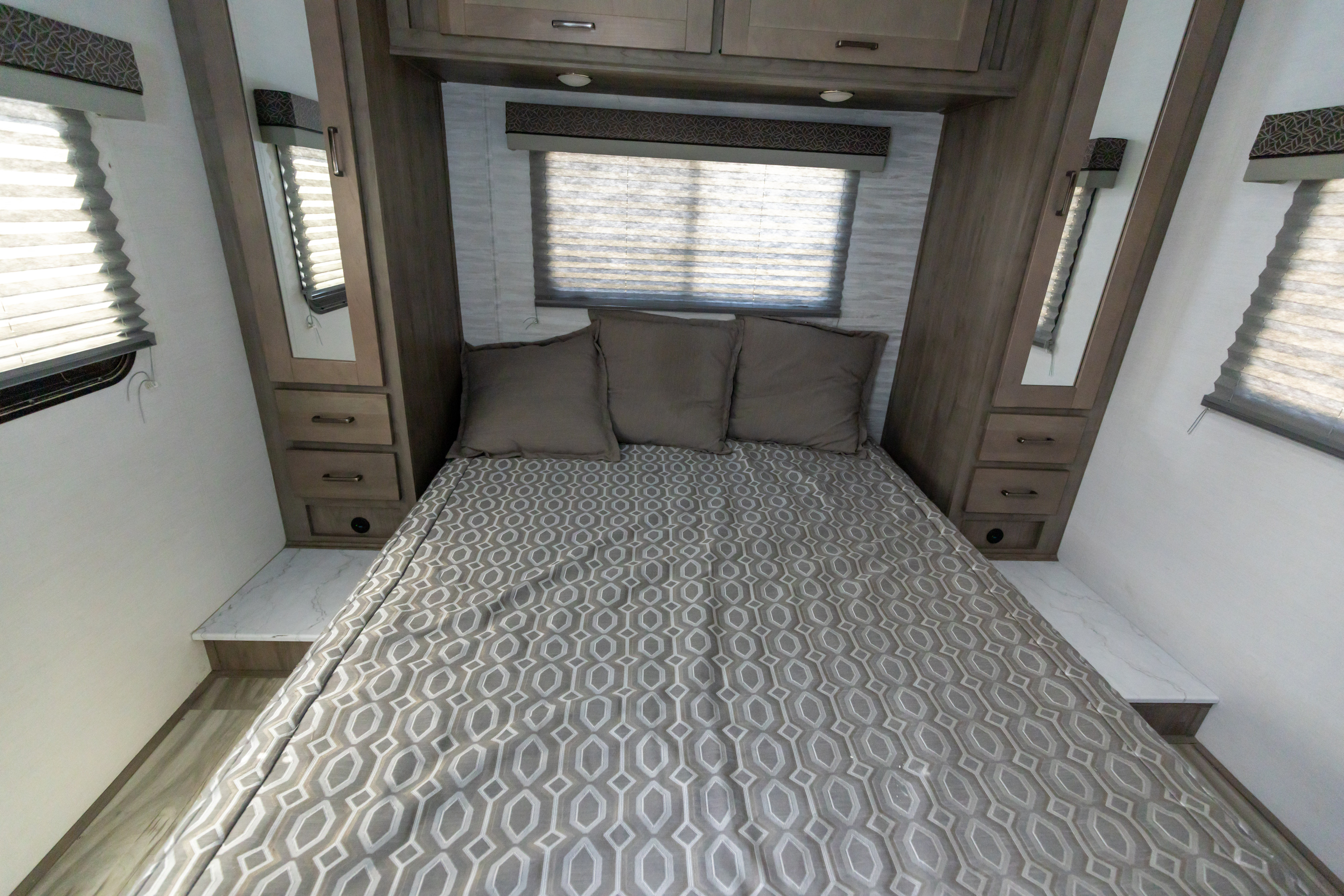 RV rental bedroom with patterned bed, three pillows, overhead cabinets, side storage, and a window with blinds.. Gulf Stream Conquest 2024