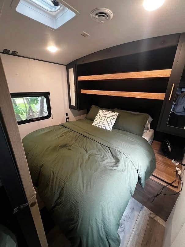 Cozy RV rental bedroom with olive green bedding, a modern headboard, and natural light from the window.. Keystone RV Springdale 2023