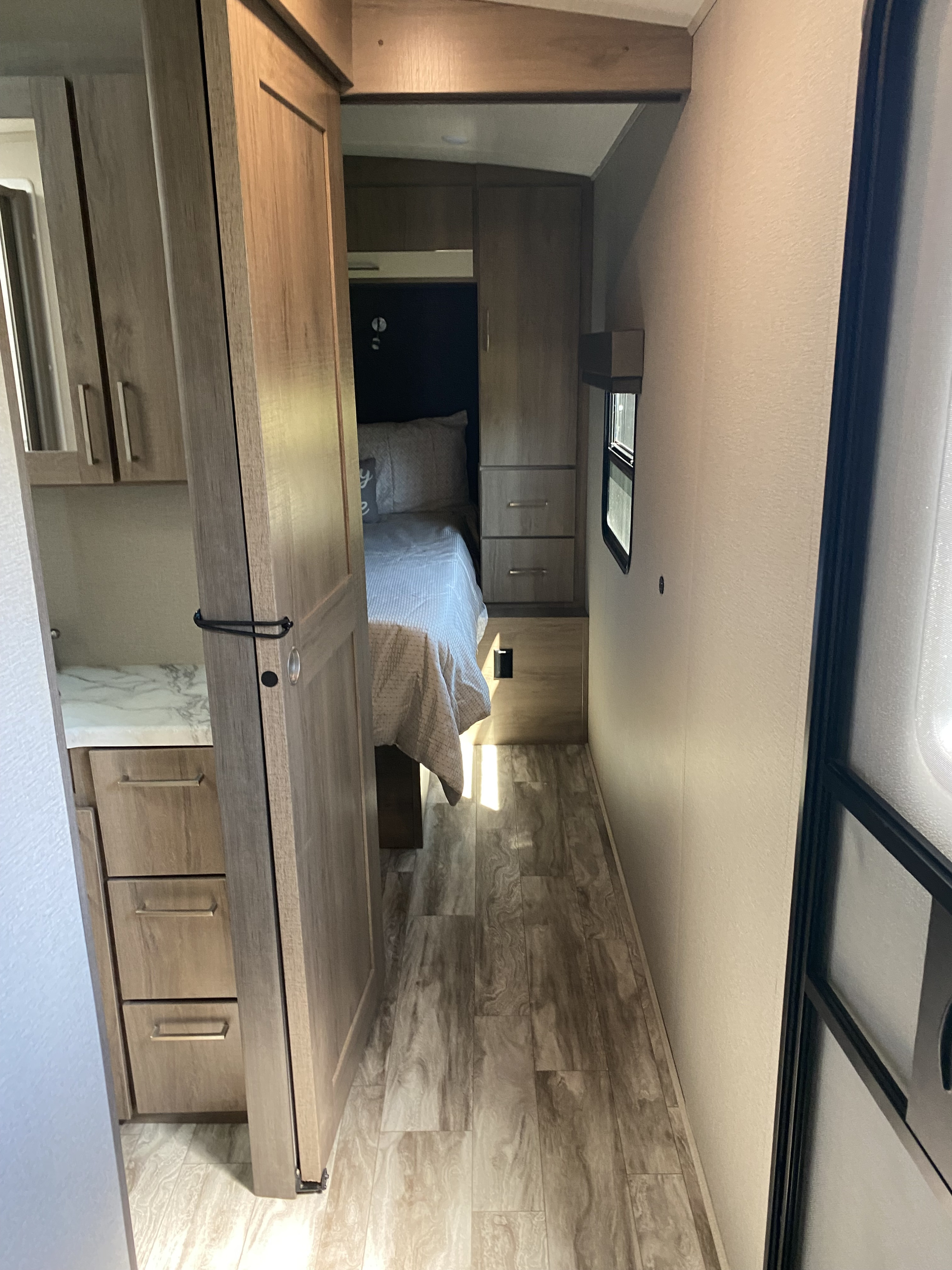 Narrow RV rental hallway leads to a cozy bedroom with bed, built-in cabinets, and a side window.. Grand Design Imagine 2021