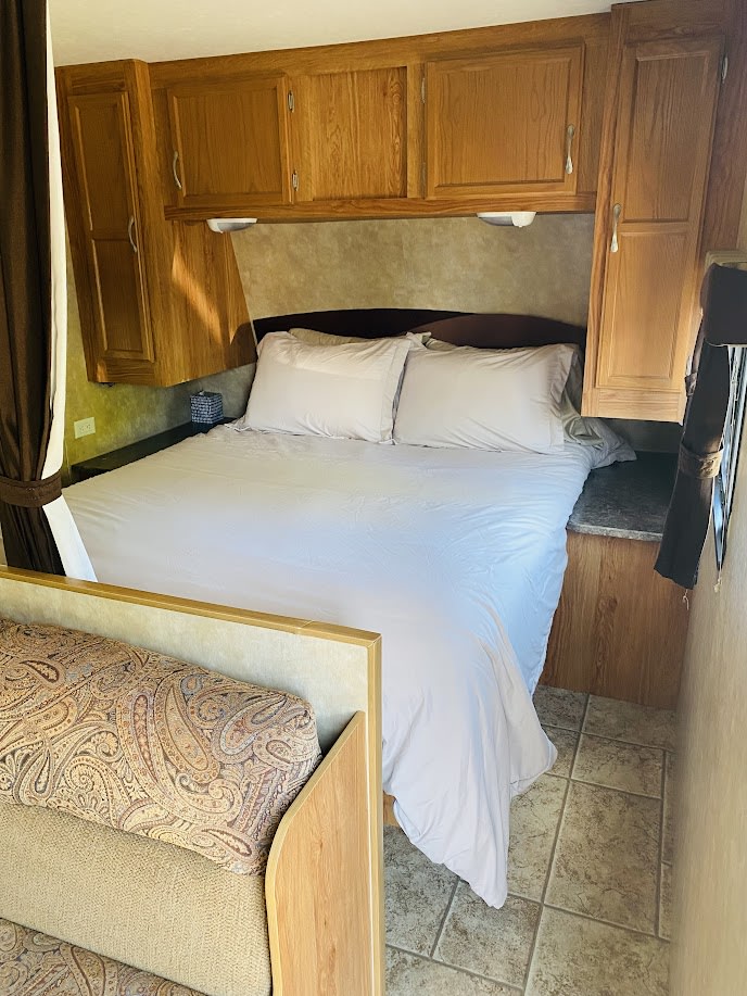 RV rental bedroom: A comfortable bed with white linens, wood cabinets, and tiled floor, adjacent to a patterned sofa.. Jayco Jay Flight 2010