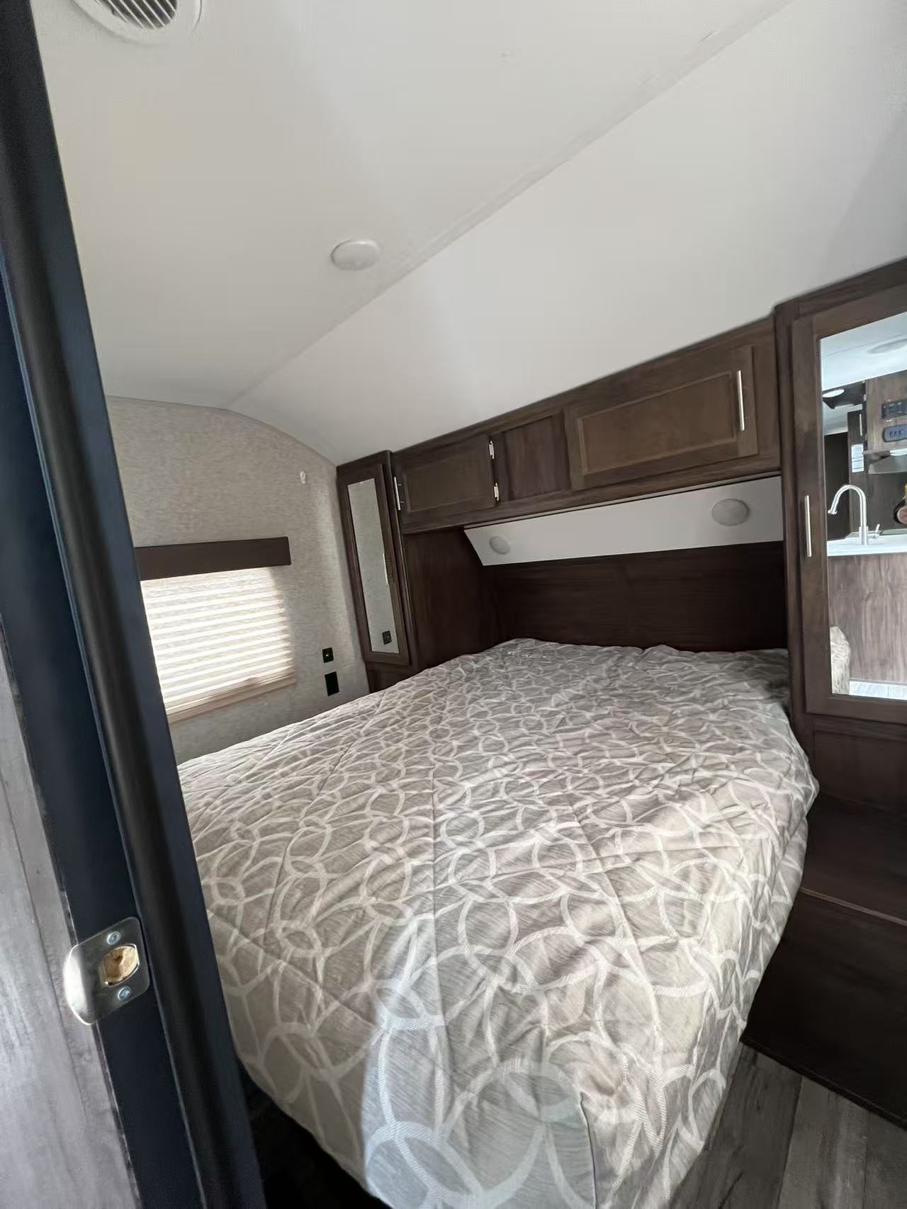 Cozy RV rental bedroom featuring a large bed with patterned comforter, overhead storage cabinets, and a window with blinds.. Forest River Cherokee 2018