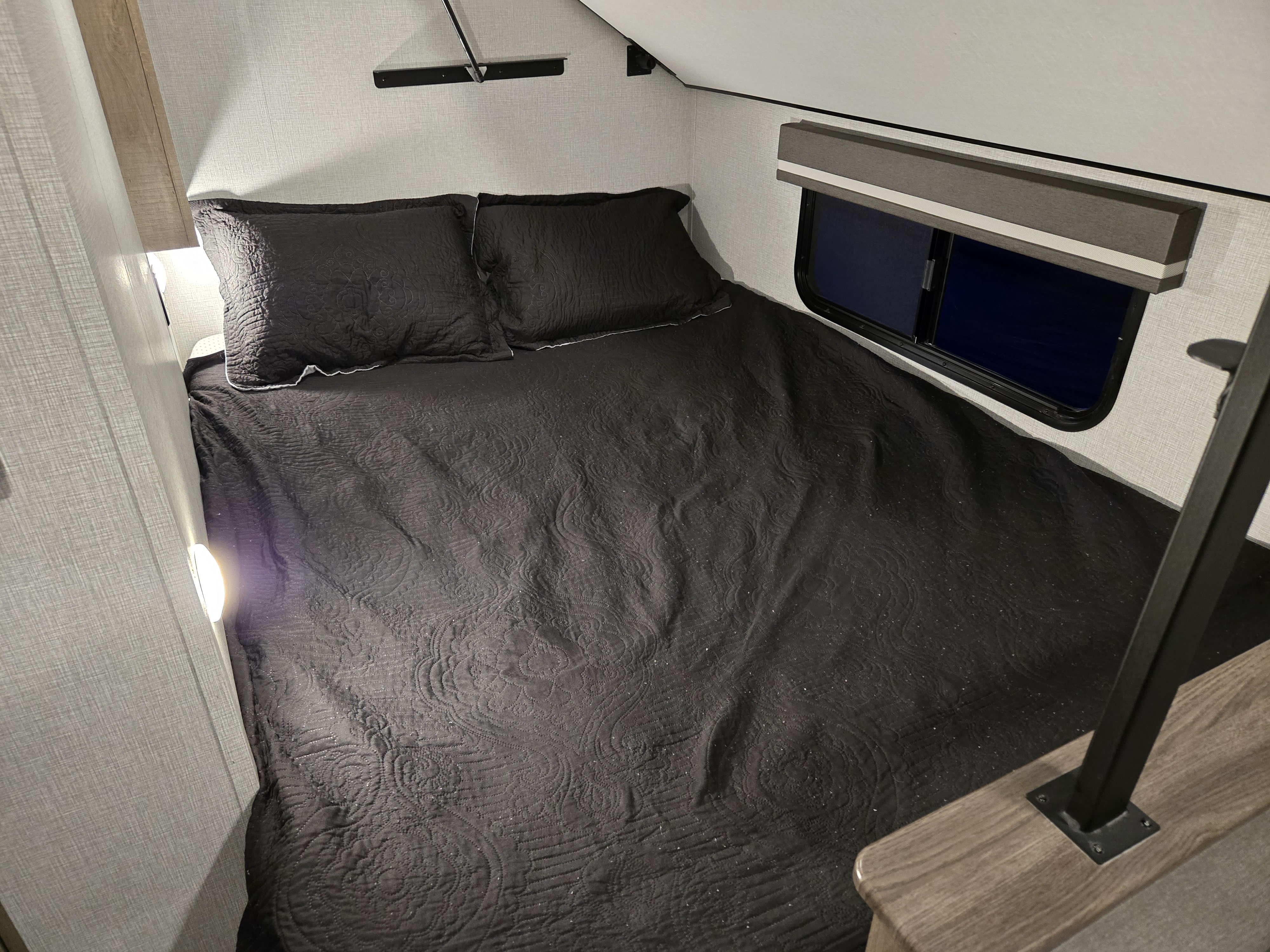 Inside an RV rental, discover a compact bedroom space with a dark quilted bed, small window, and a wall-mounted reading light.. Grand Design Imagine AIM 2023