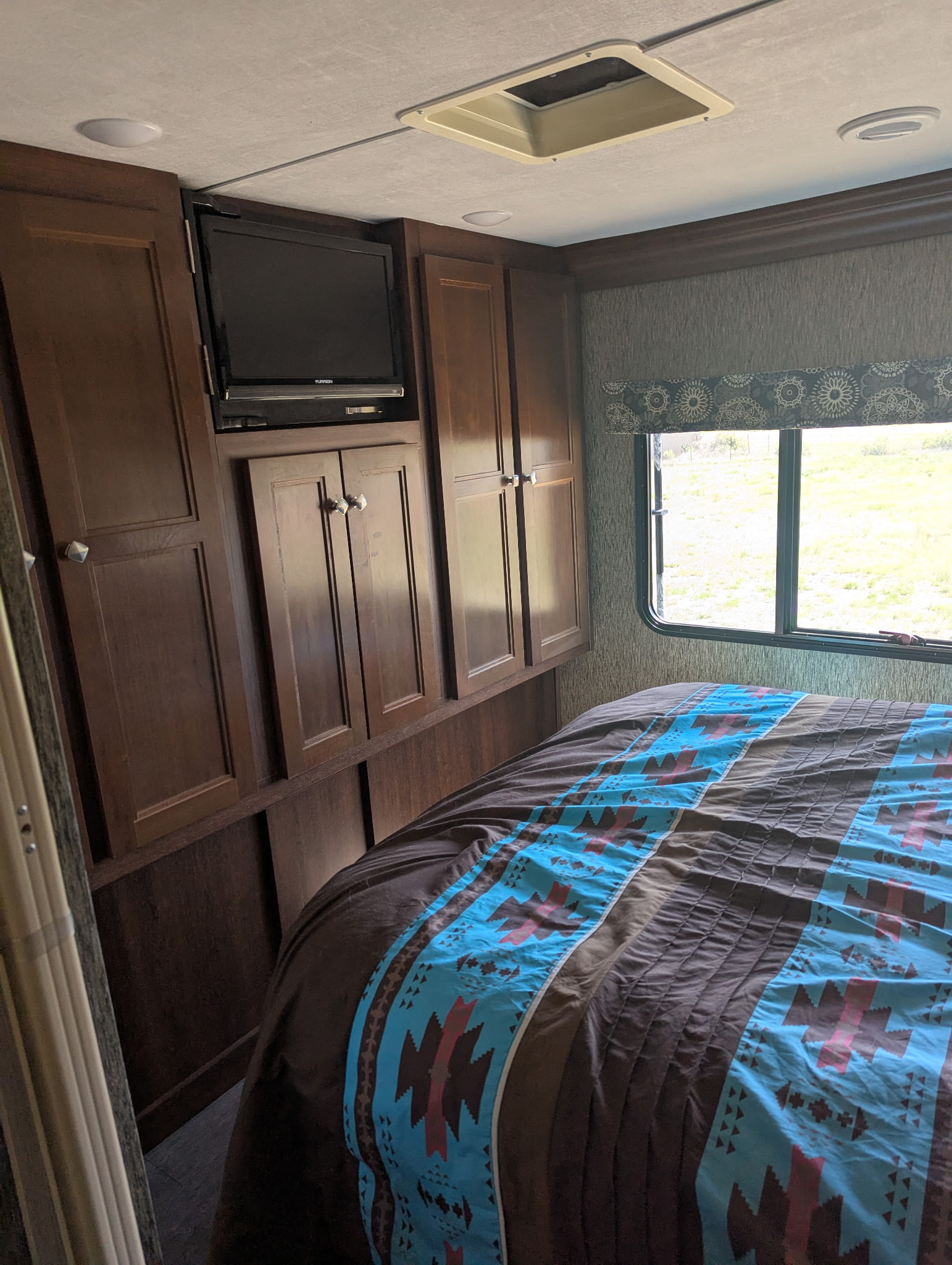 Inside an RV rental, this bedroom features a bed with a patterned comforter, built-in wood cabinets with a TV, and a window view.. Forest River Sunseeker 2017