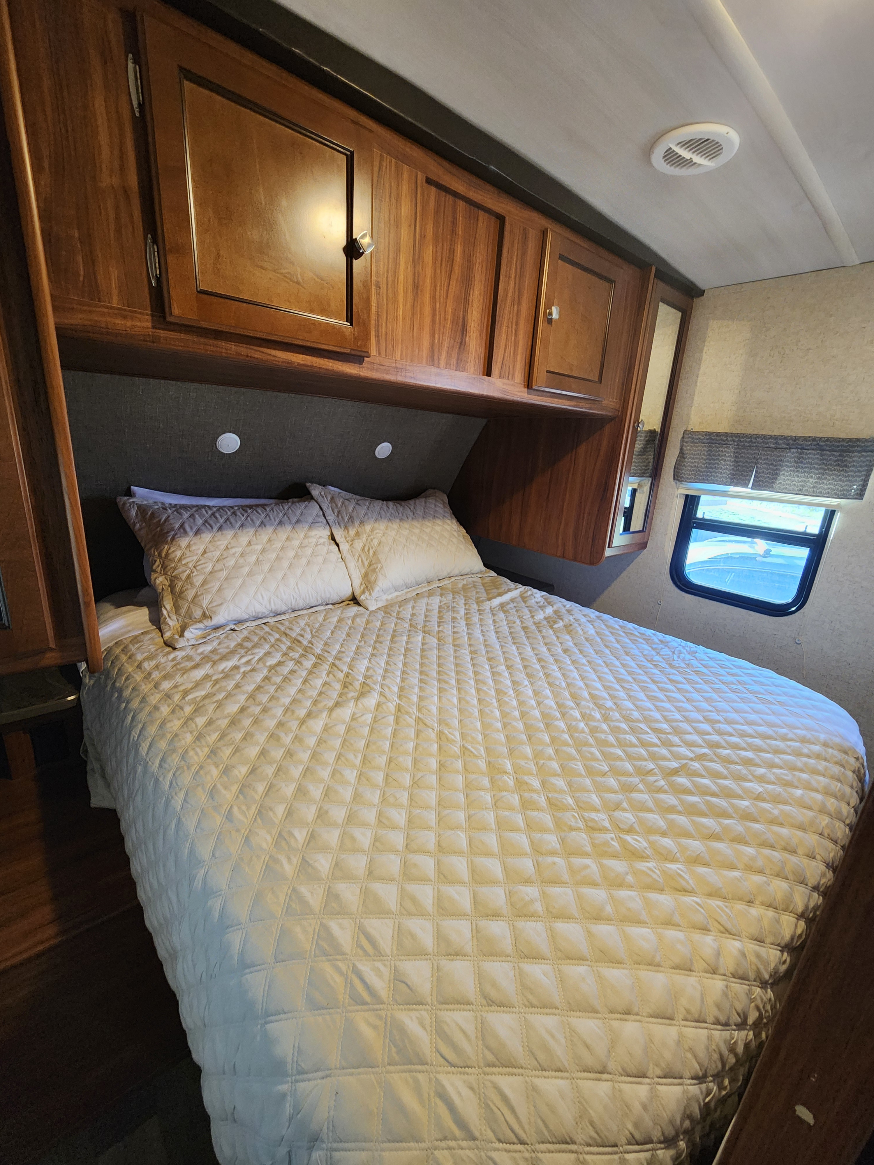 Cozy RV rental bedroom featuring a quilted bed, wooden overhead cabinets, and a side window.. Heartland RVs Mallard 2017
