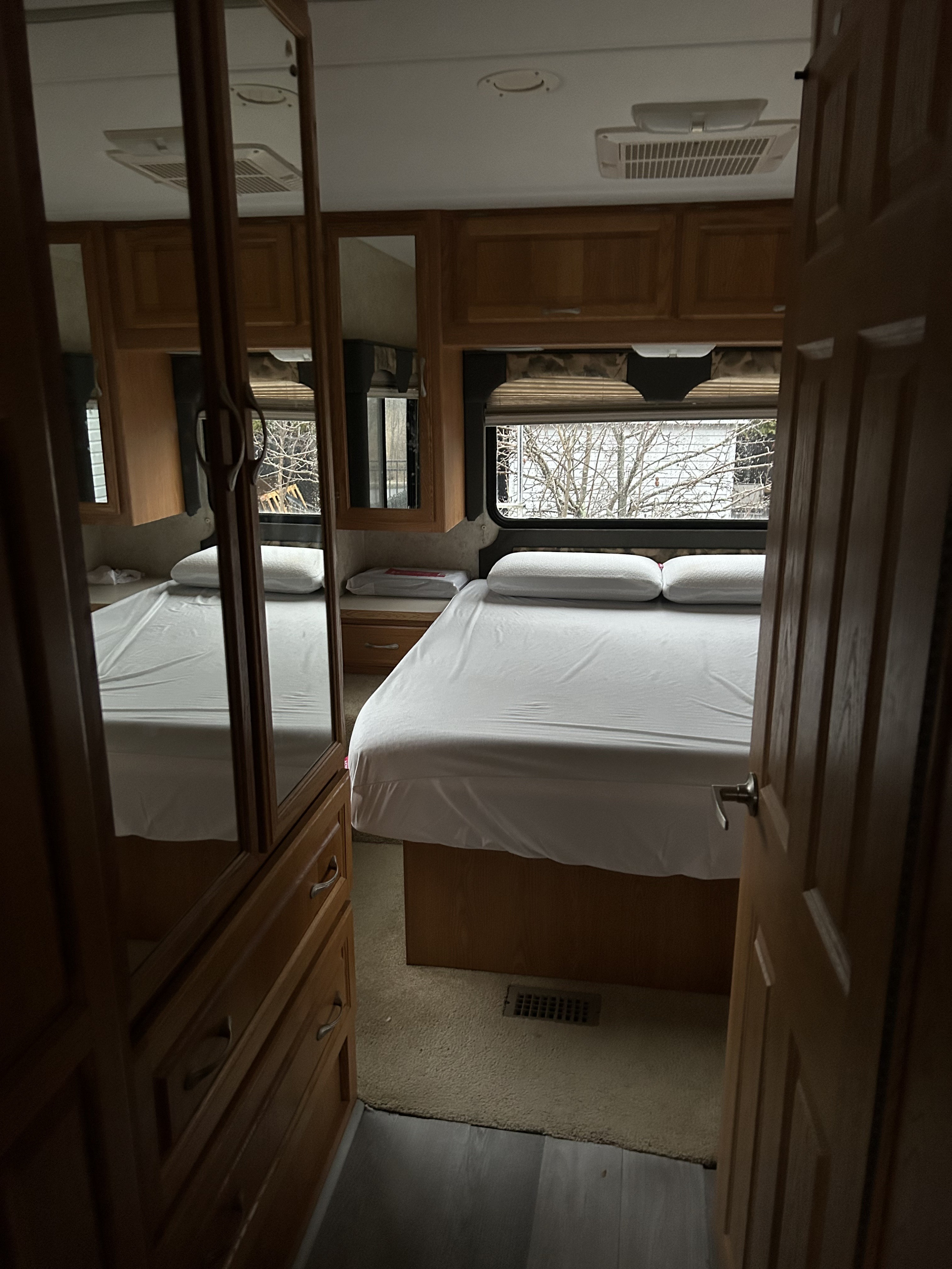 Cozy RV rental bedroom showing a white bed, overhead cabinets, window view, and mirrored closet on left.. Four Winds Hurricane 2006