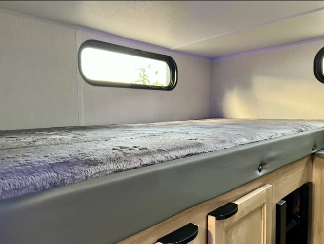 Inside an RV rental: a cozy bunk bed with a soft grey blanket, oval windows, and light wood cabinets below.. Forest River cherokee Grey Wolf 2024