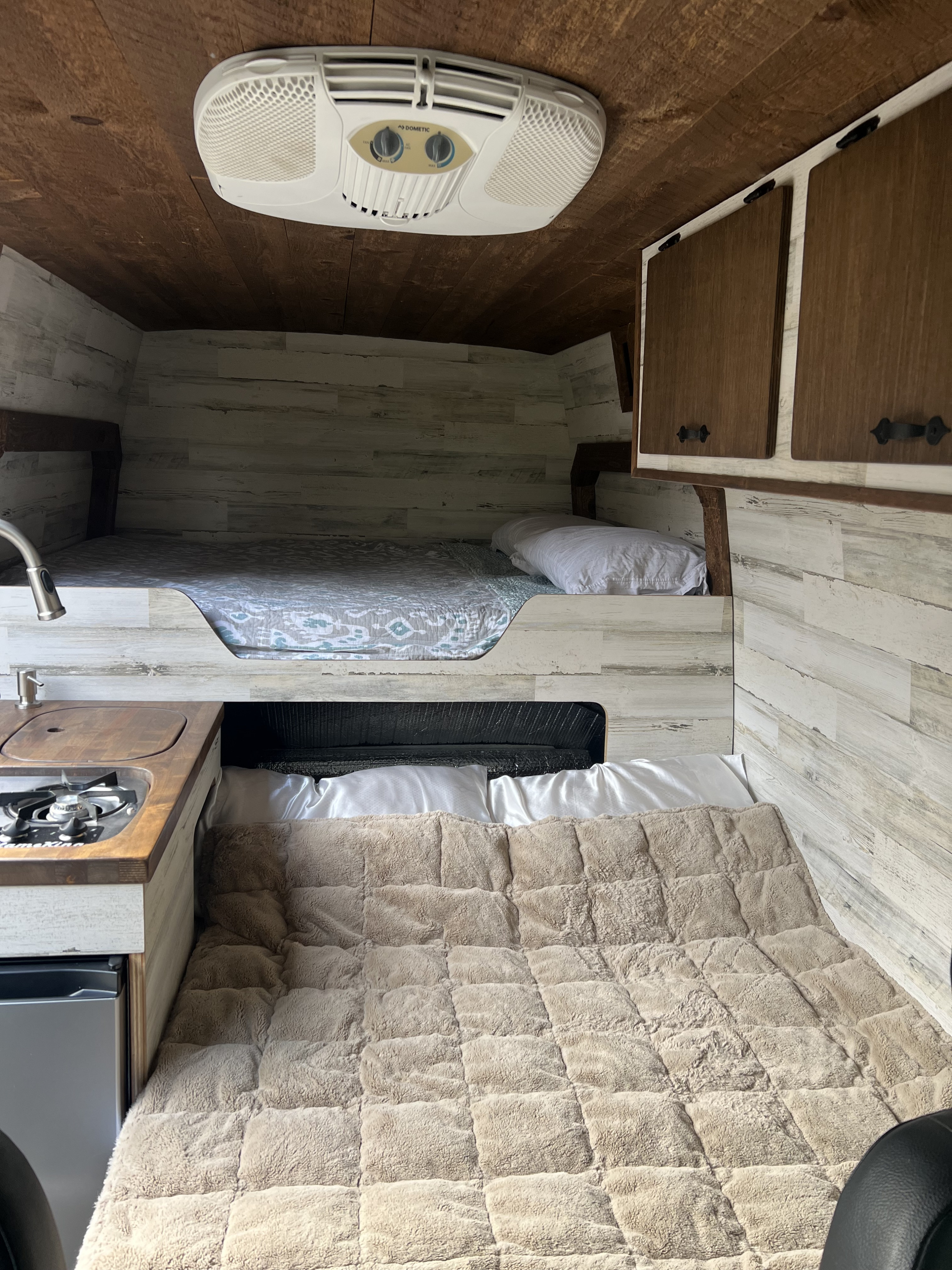 Inside an RV rental: two comfy bunk beds, compact kitchen with fridge, and AC unit. Walls feature distressed white wood paneling.. Mercedes-Benz sprinter 2019