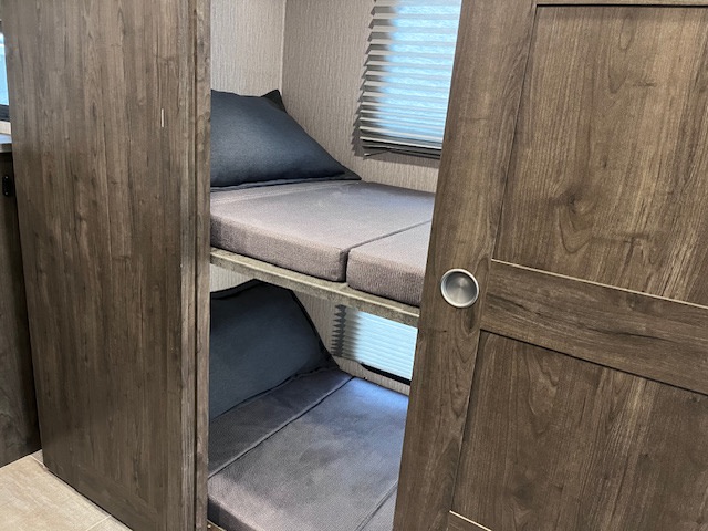 This RV rental interior shows a two-tier bunk bed with grey cushions, a window with blinds, and a wooden door on the right.. Forest River Coachmen Mirada 2022