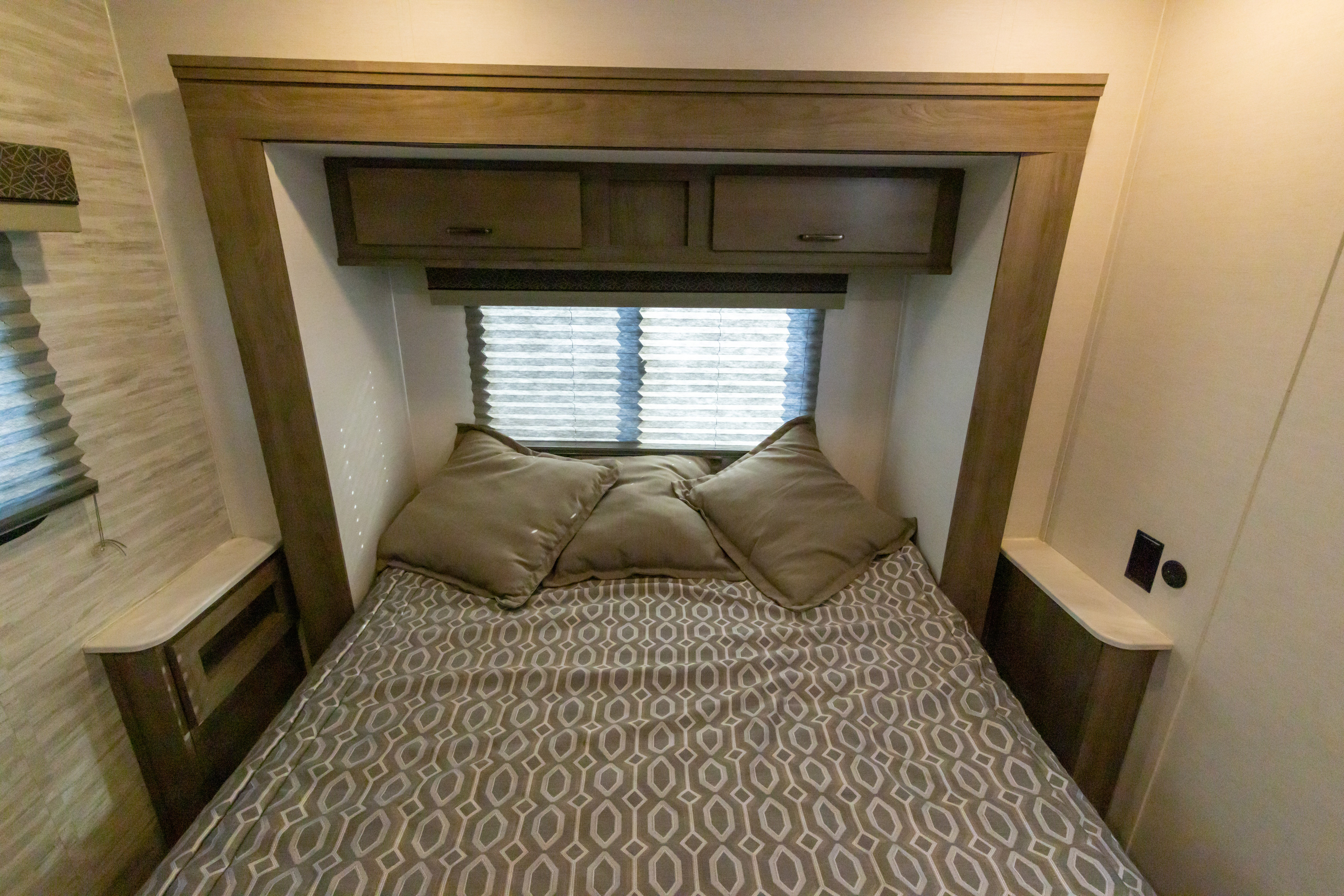 Compact RV rental bedroom features a bed with patterned bedding, pillows, overhead cabinets, and side shelves. Window with blinds.. Gulf Stream Conquest 2024