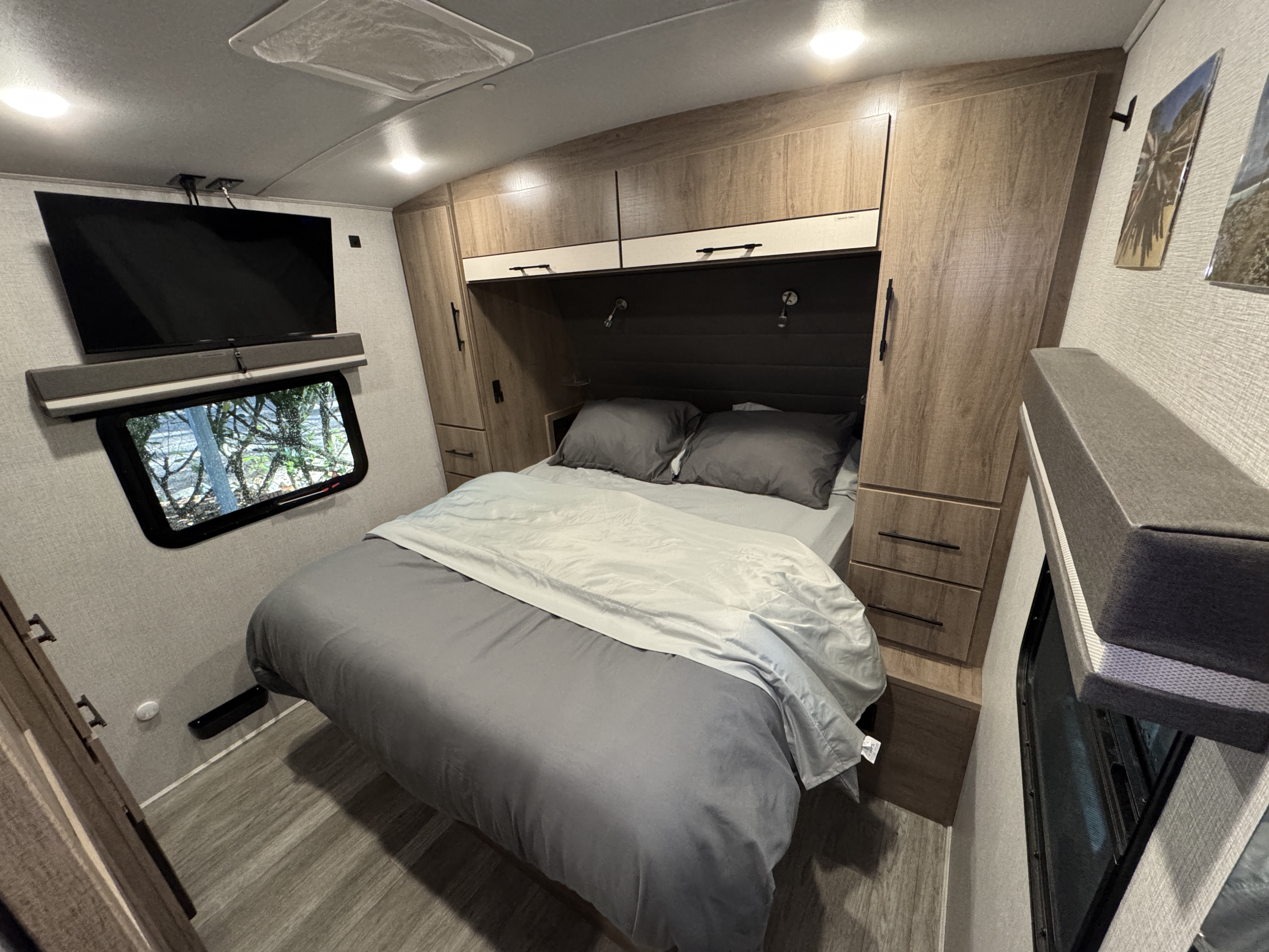 Experience comfort in this RV rental bedroom with a cozy bed, ample storage, wall-mounted TV, and window views.. Grand Design Imagine 2023