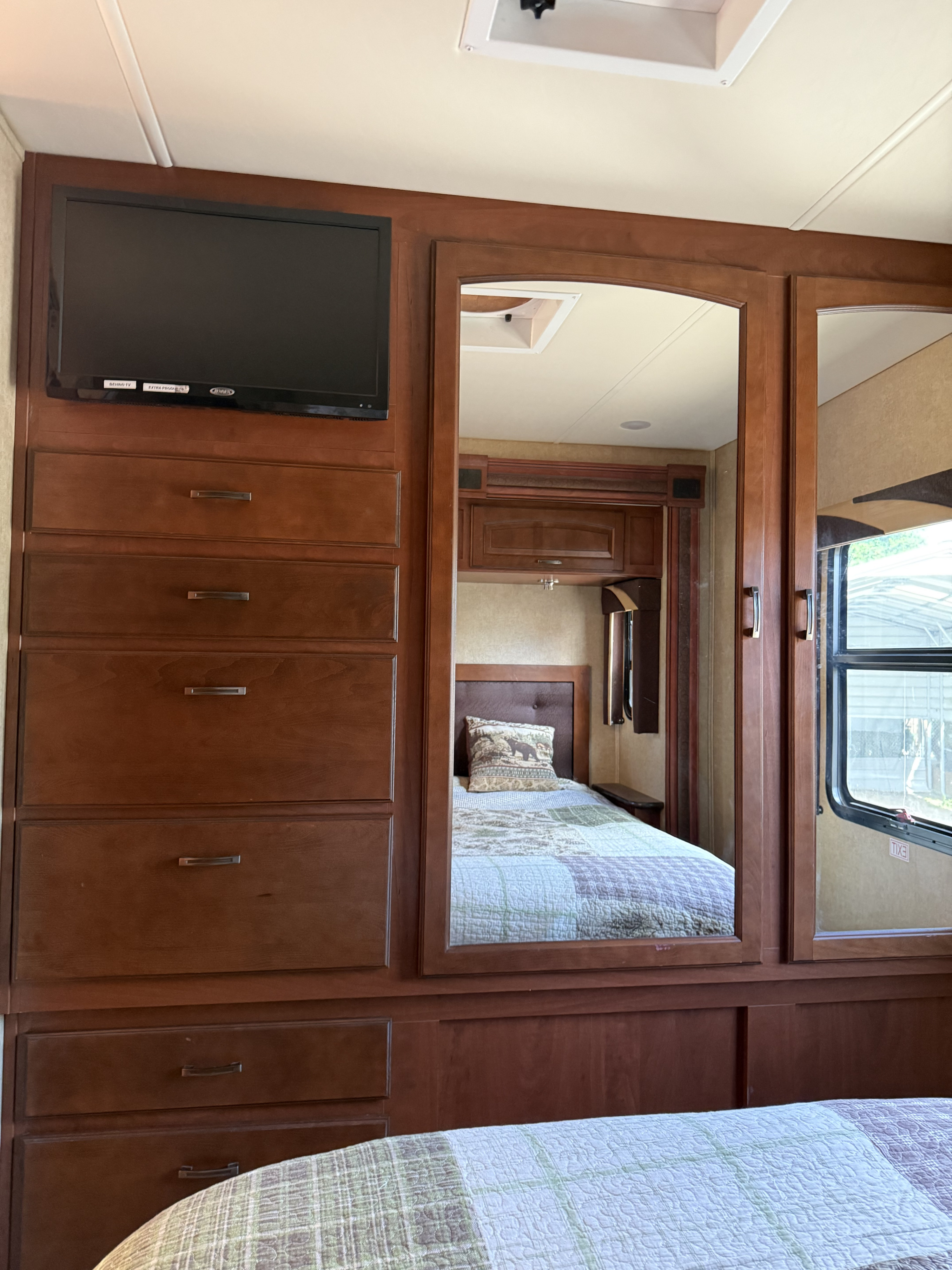 Inside an RV rental: a wooden dresser with TV, built-in storage, and mirror reflecting a cozy bedroom with patterned quilt.. Jayco Greyhawk 2015