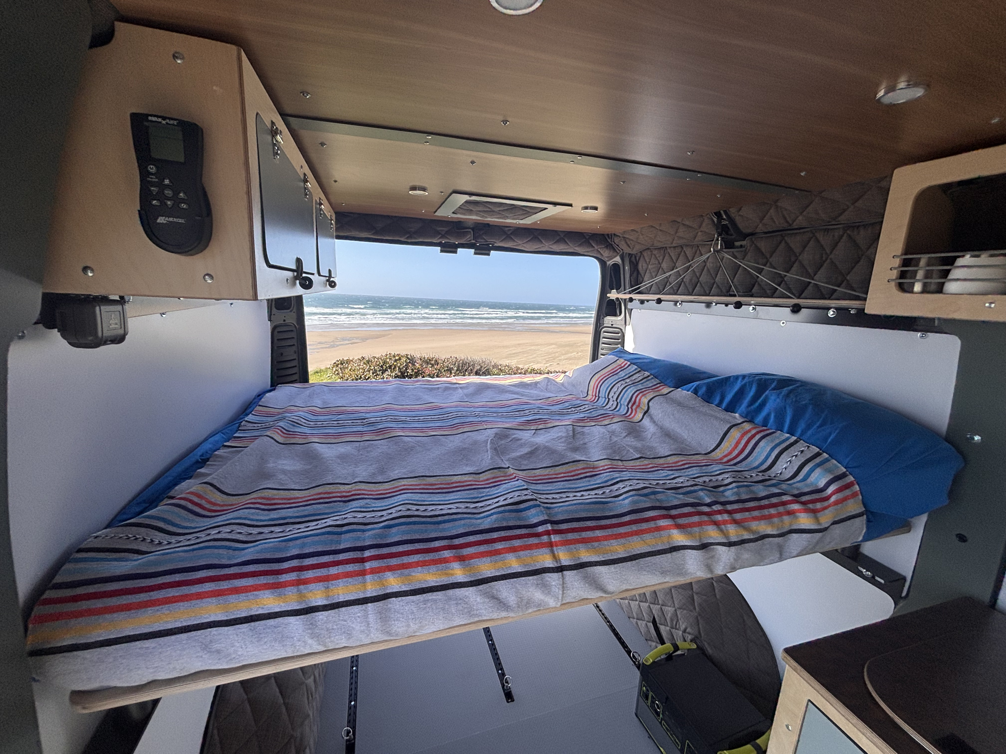 Inside a comfortable RV rental, a bed faces a stunning ocean view, offering a serene travel experience.. RAM RAM PROMASTER 1500 2024
