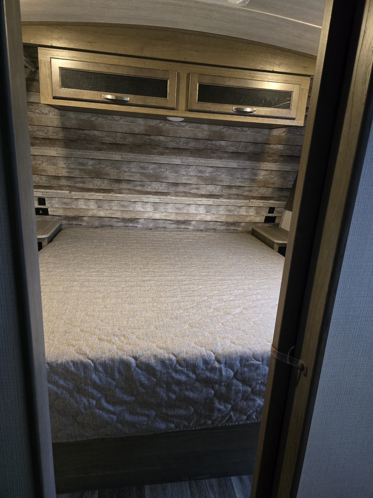Inside an RV rental, a rustic bedroom features a comfortable quilted bed, wood-paneled wall, and overhead cabinets.. Keystone RV Cougar 2021